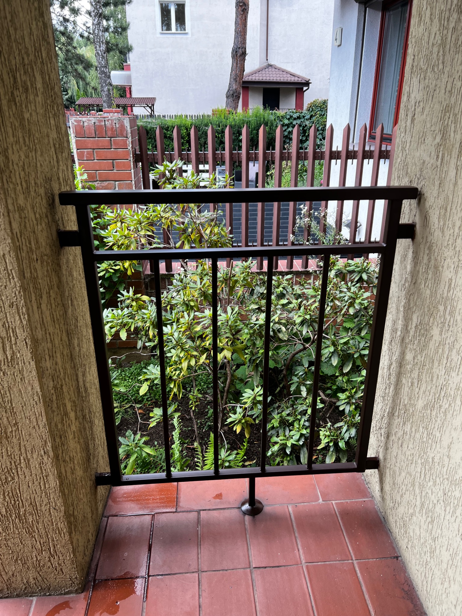 Ciemna, metalowa balustrada balkonowa z widokiem na bujną zieleń ogrodu, widziana z zewnątrz, z mokrą, czerwoną terakotą na podłodze.
