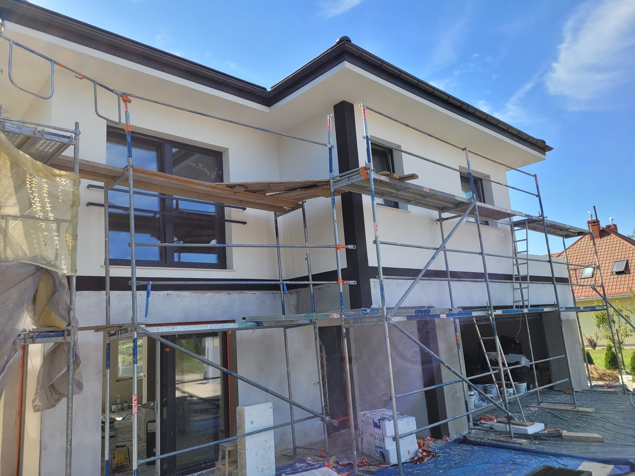 Dom jednorodzinny w trakcie prac elewacyjnych, widoczne rusztowania, świeżo pomalowana biała elewacja z ciemnymi akcentami, przygotowane materiały budowlane i zabezpieczenia.