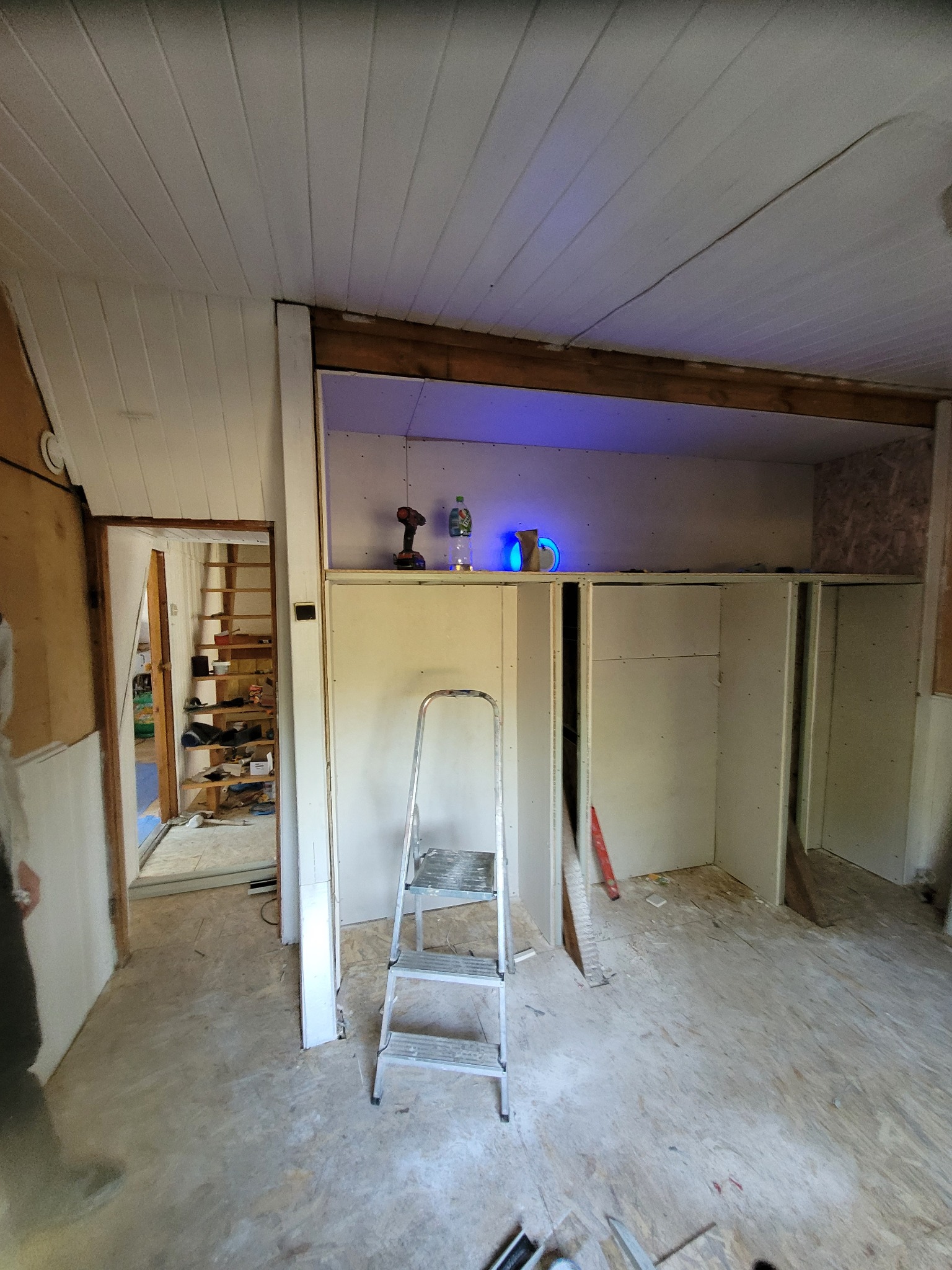 Wnętrze w trakcie prac wykończeniowych: widoczne płyty kartonowo-gipsowe tworzące zabudowę, drabina aluminiowa, narzędzia i akcesoria na półce z podświetleniem LED, oraz otwarte przejście do innego...