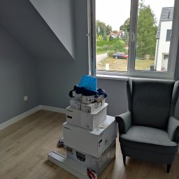 Viacheslav Holovko - Pokój po remoncie, z szarą ścianą i podłogą z paneli, z stertą pudeł z lampami i fotelem uszak w rogu, widok z okna na zewnątrz.