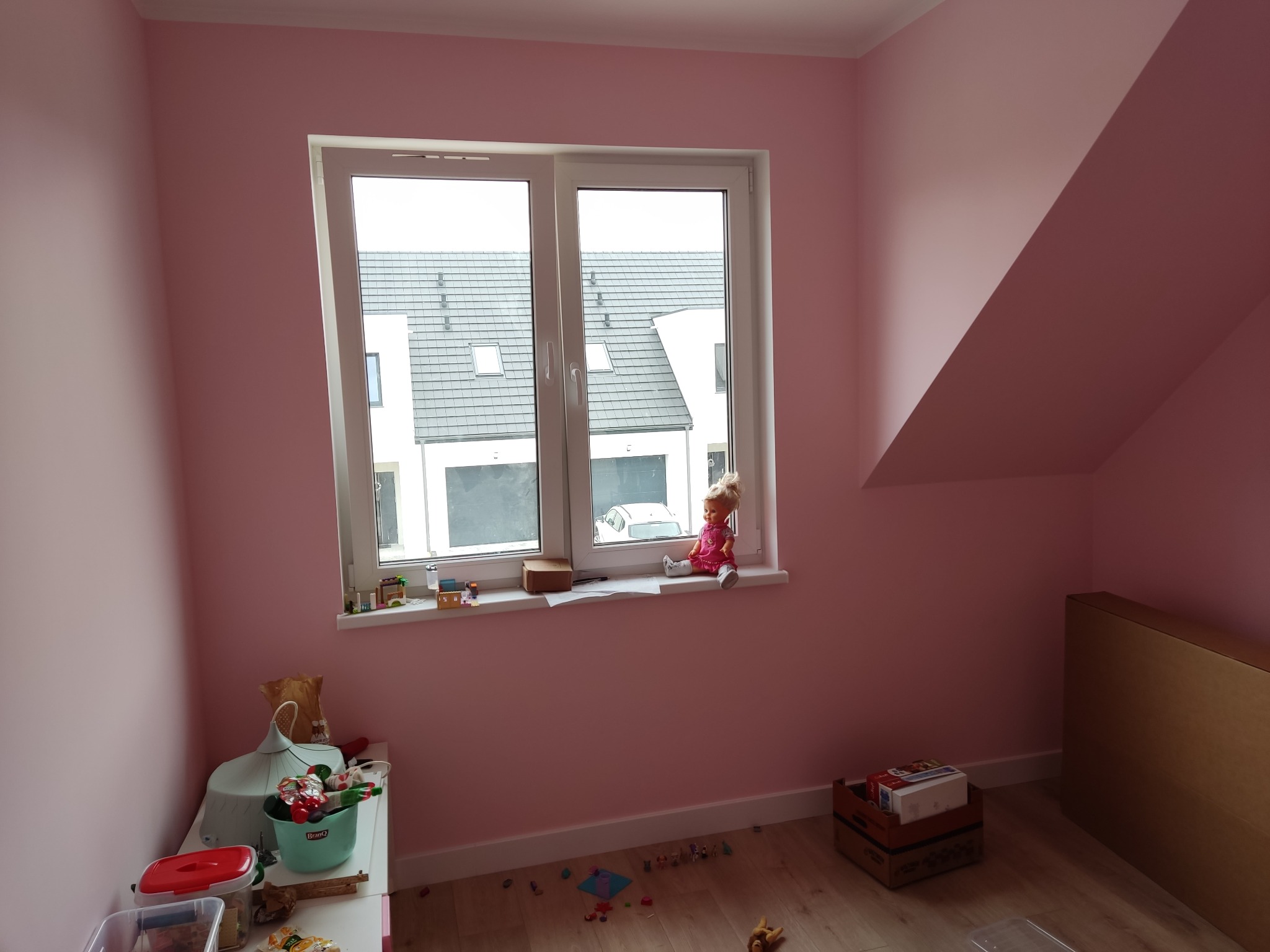 Pokój dziecięcy z pomalowanymi na różowo ścianami, białe okno z widokiem na budynki, zabawki na parapecie i podłodze, kartony przy ścianie.