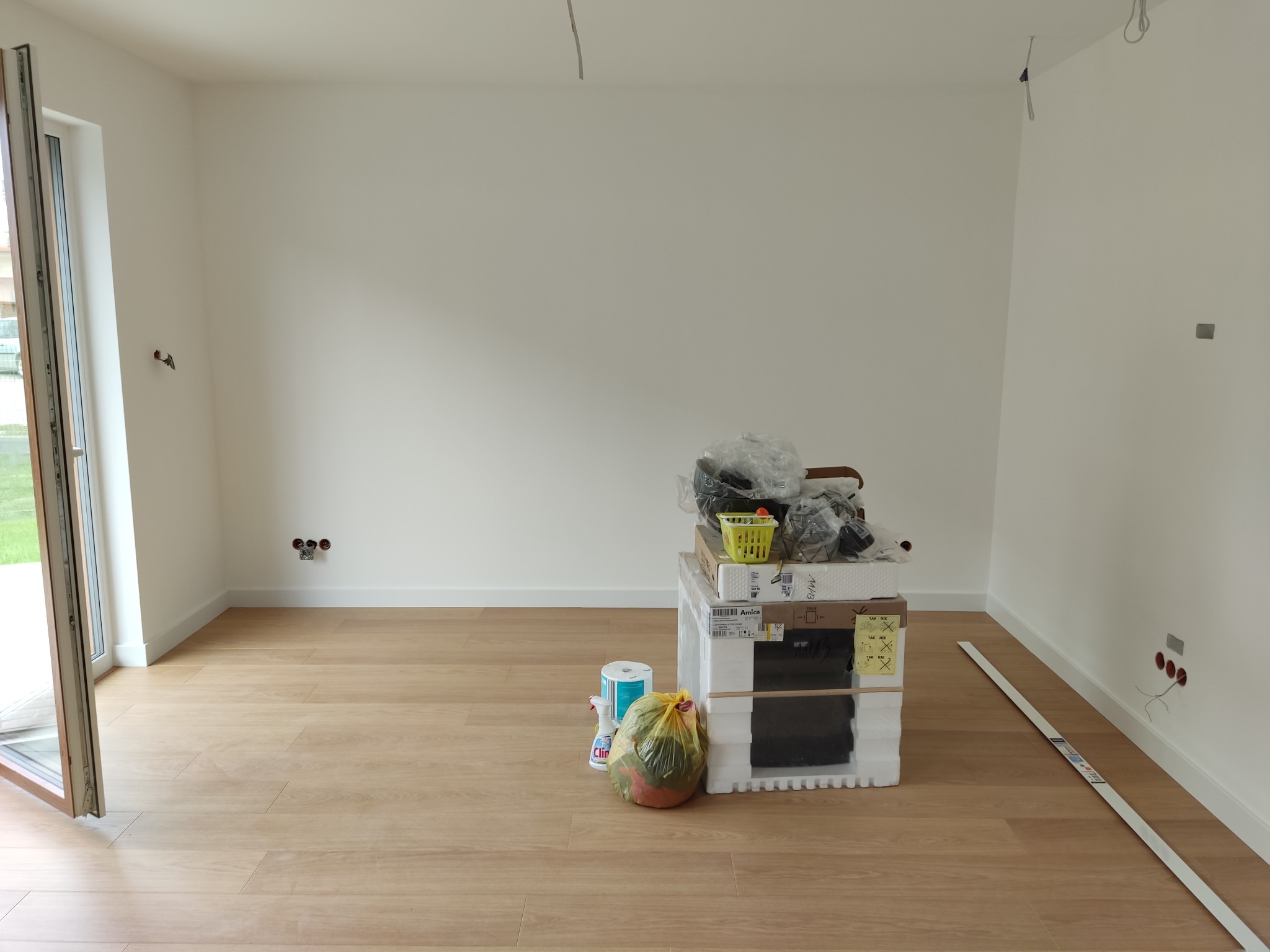 Pokój po remoncie z nową podłogą panelową, świeżo pomalowanymi ścianami, widoczne gniazdka elektryczne i listwy przypodłogowe, na środku pomieszczenia stos materiałów budowlanych i opakowań.