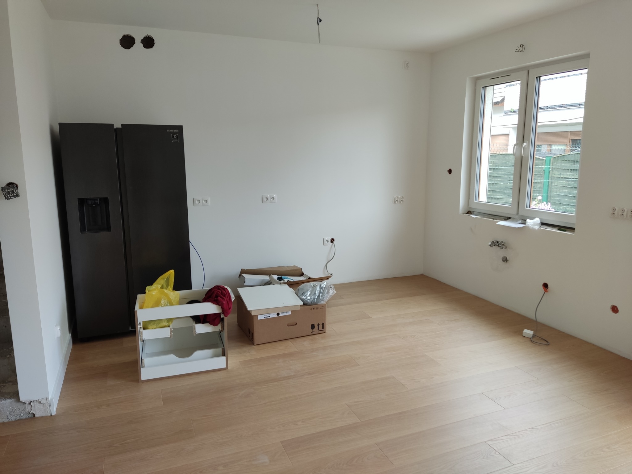 Jasne panele podłogowe w nowym, pustym pomieszczeniu z białymi ścianami, widoczne gniazdka elektryczne, otwory w ścianie i stojąca lodówka Samsung.