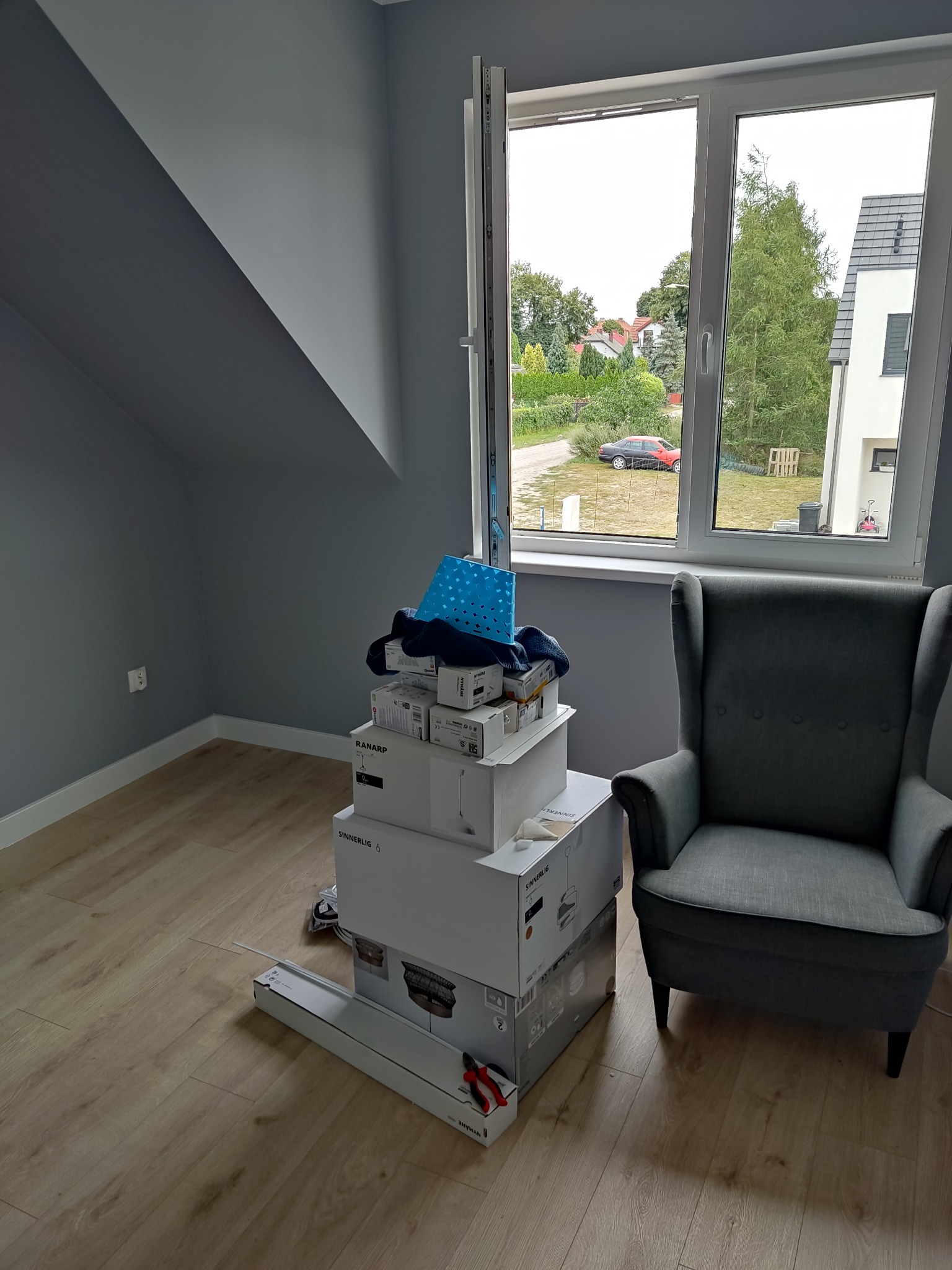 Pokój po remoncie, z szarą ścianą i podłogą z paneli, z stertą pudeł z lampami i fotelem uszak w rogu, widok z okna na zewnątrz.