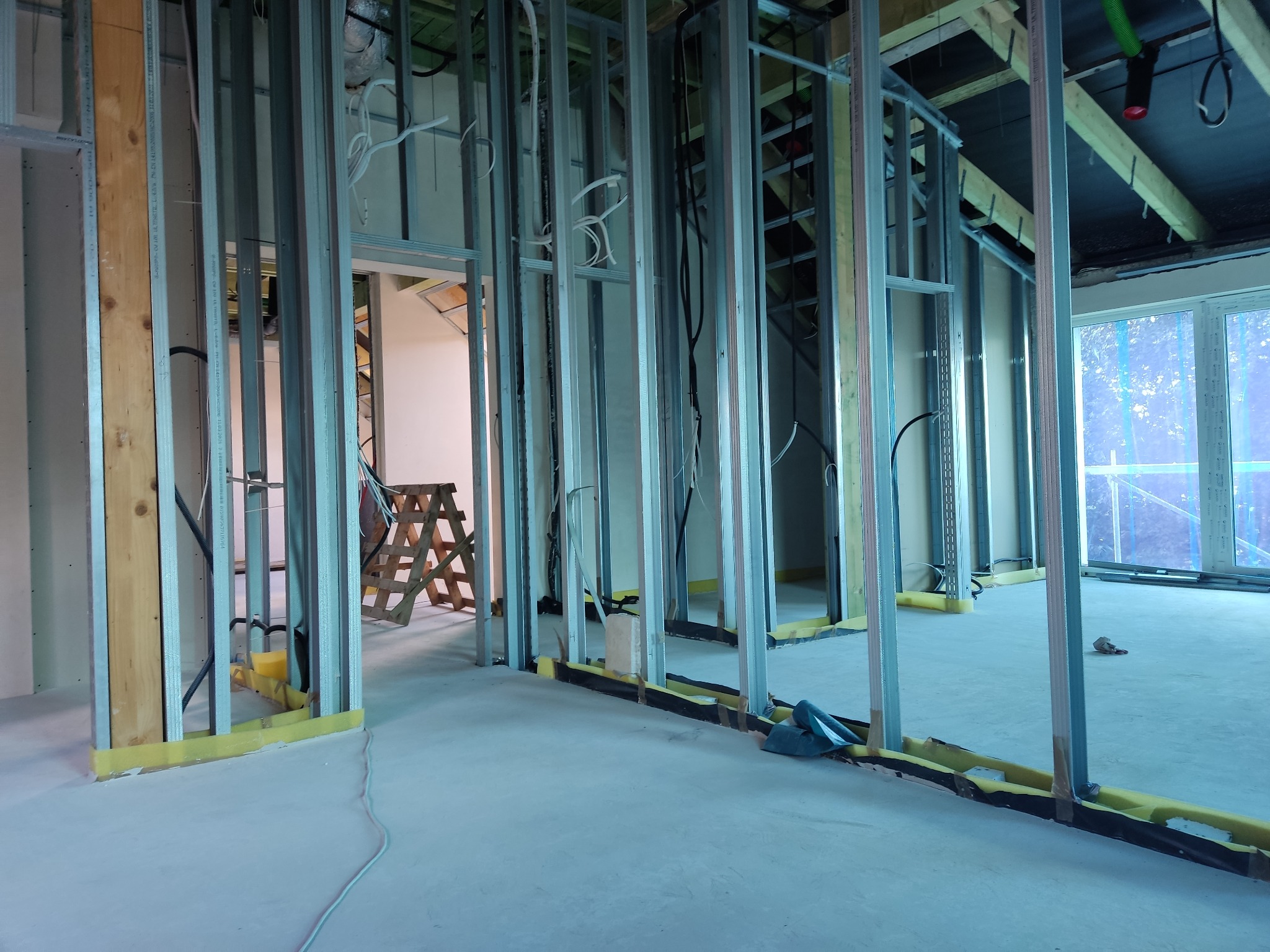Konstrukcja szkieletowa z profili stalowych wewnątrz budynku, widoczne przewody, drabina drewniana w tle, żółta taśma izolacyjna na podłodze, okno w głębi pomieszczenia.