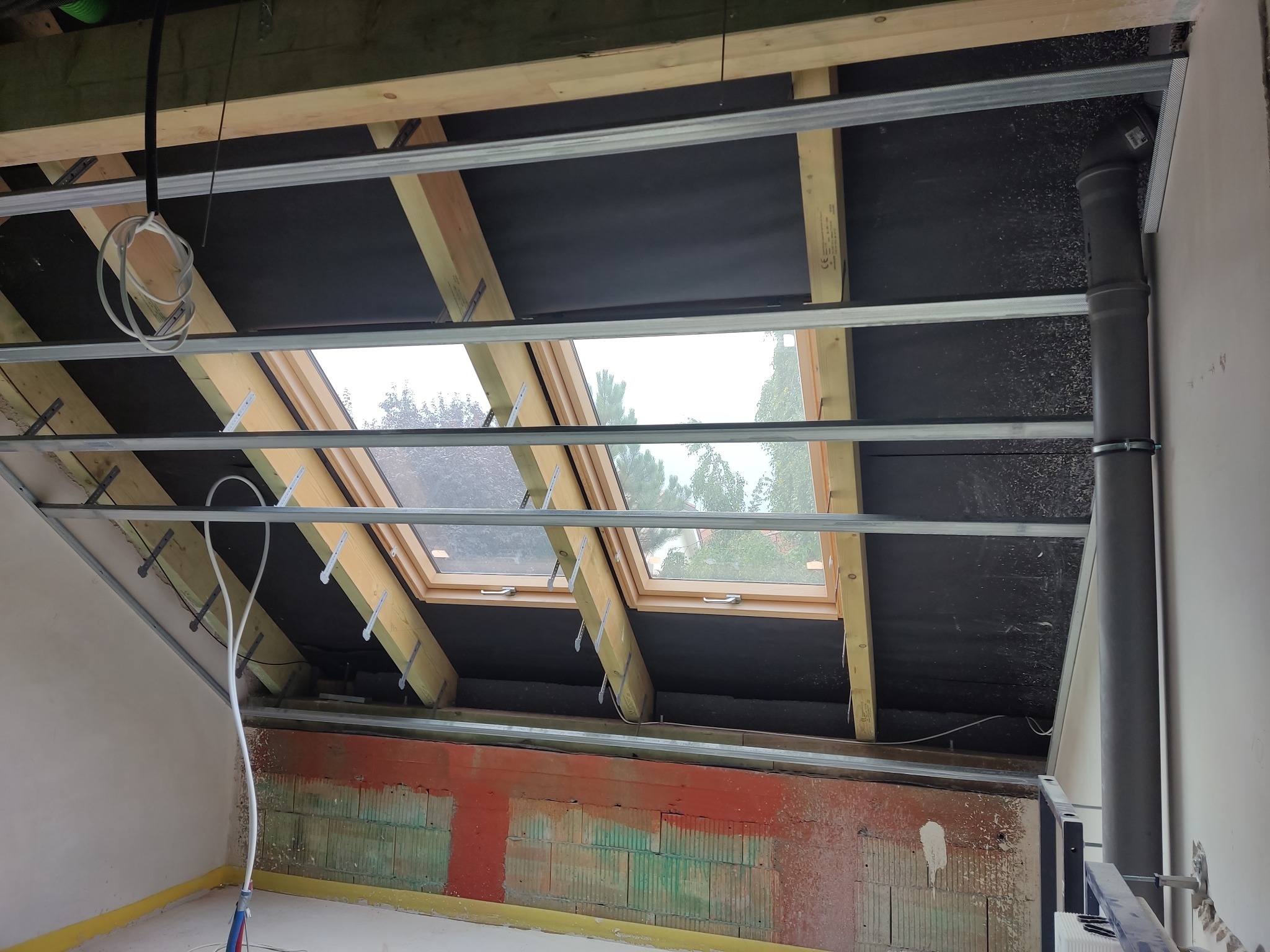 Montaż metalowego stelażu pod płytę gipsowo-kartonową na poddaszu, widoczne okna dachowe, izolacja czarną folią i fragmenty instalacji hydraulicznej.