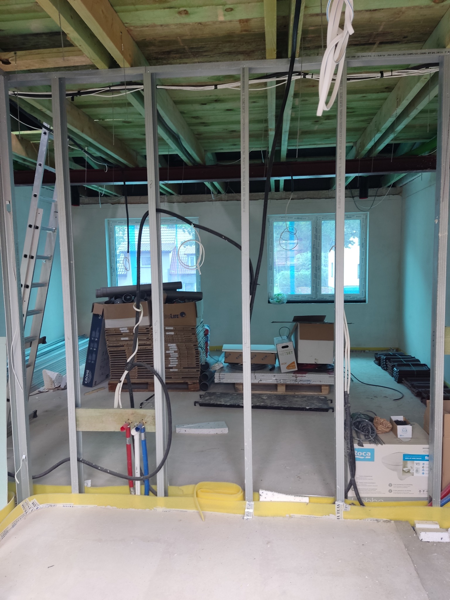 Wnętrze w trakcie prac remontowych: widoczne metalowe profile konstrukcyjne, instalacje elektryczne i hydrauliczne, materiały budowlane w kartonach, surowy strop z drewnianych belek.