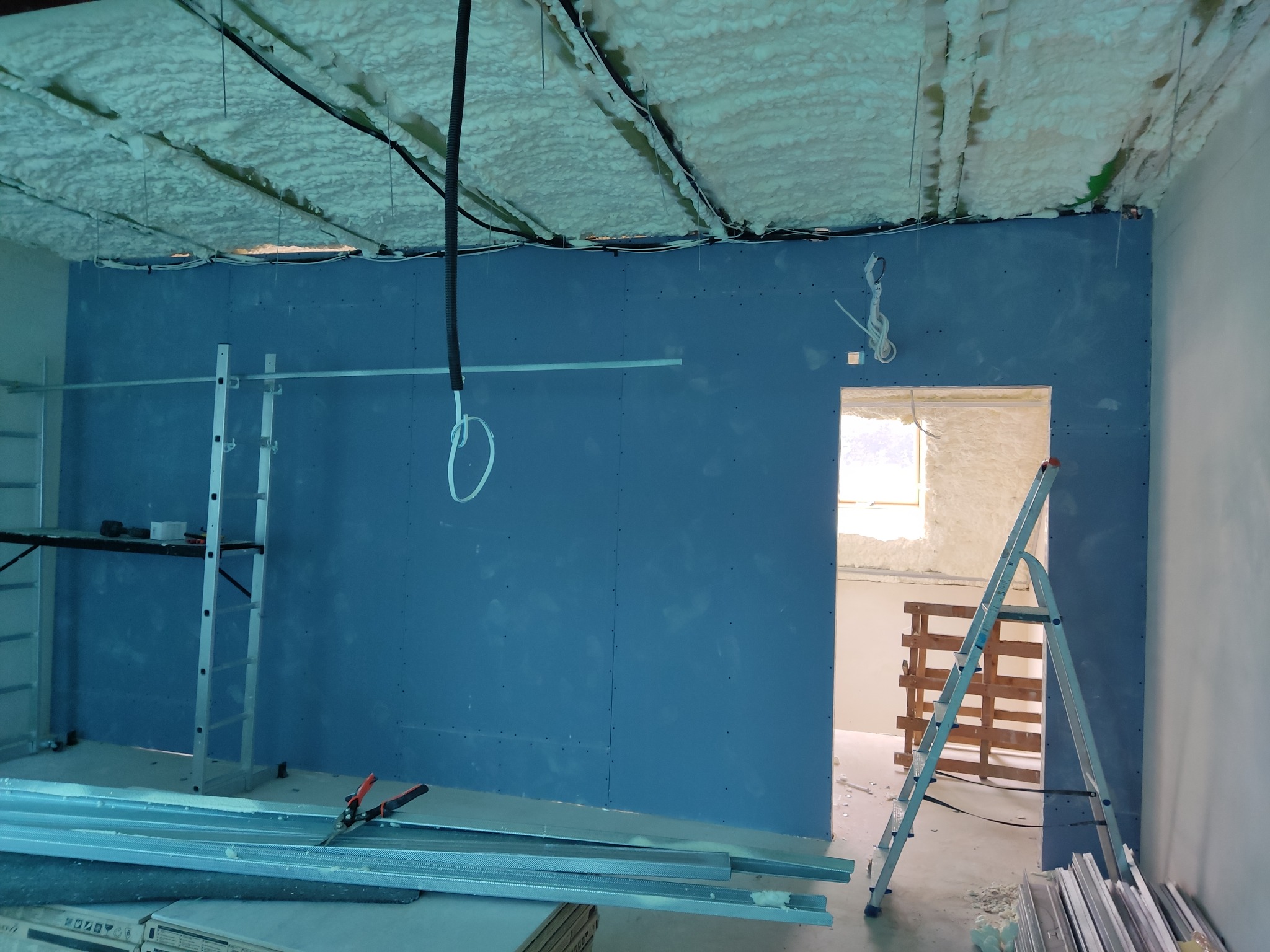 Montaż płyt kartonowo-gipsowych na ścianie w nowym budynku, widoczne profile, drabiny i piana izolacyjna na suficie.