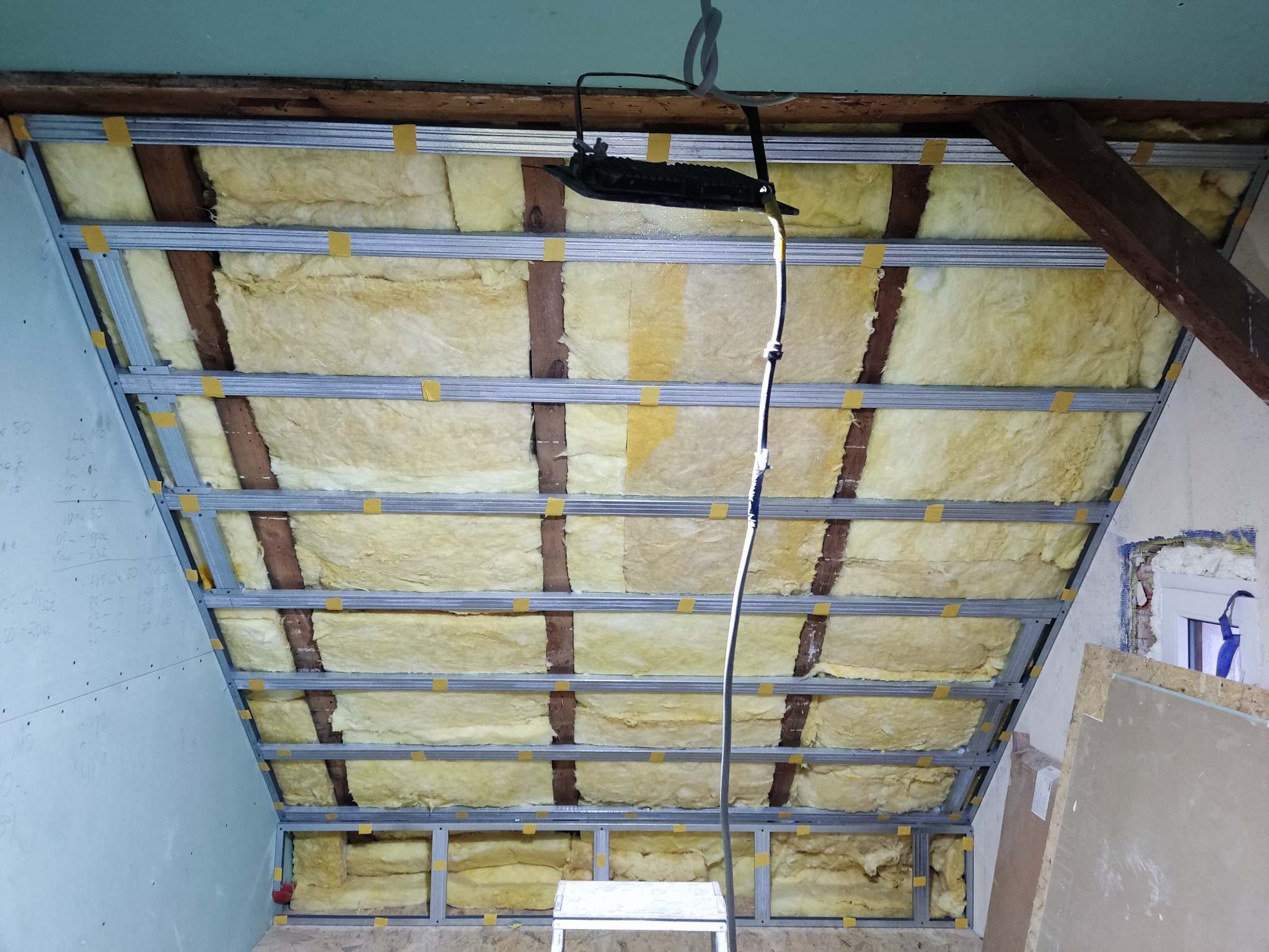 Montaż izolacji z wełny mineralnej w metalowym stelażu poddasza, zabezpieczonej żółtą taśmą, widoczna częściowo zamontowana płyta gipsowo-kartonowa z odręcznymi notatkami.