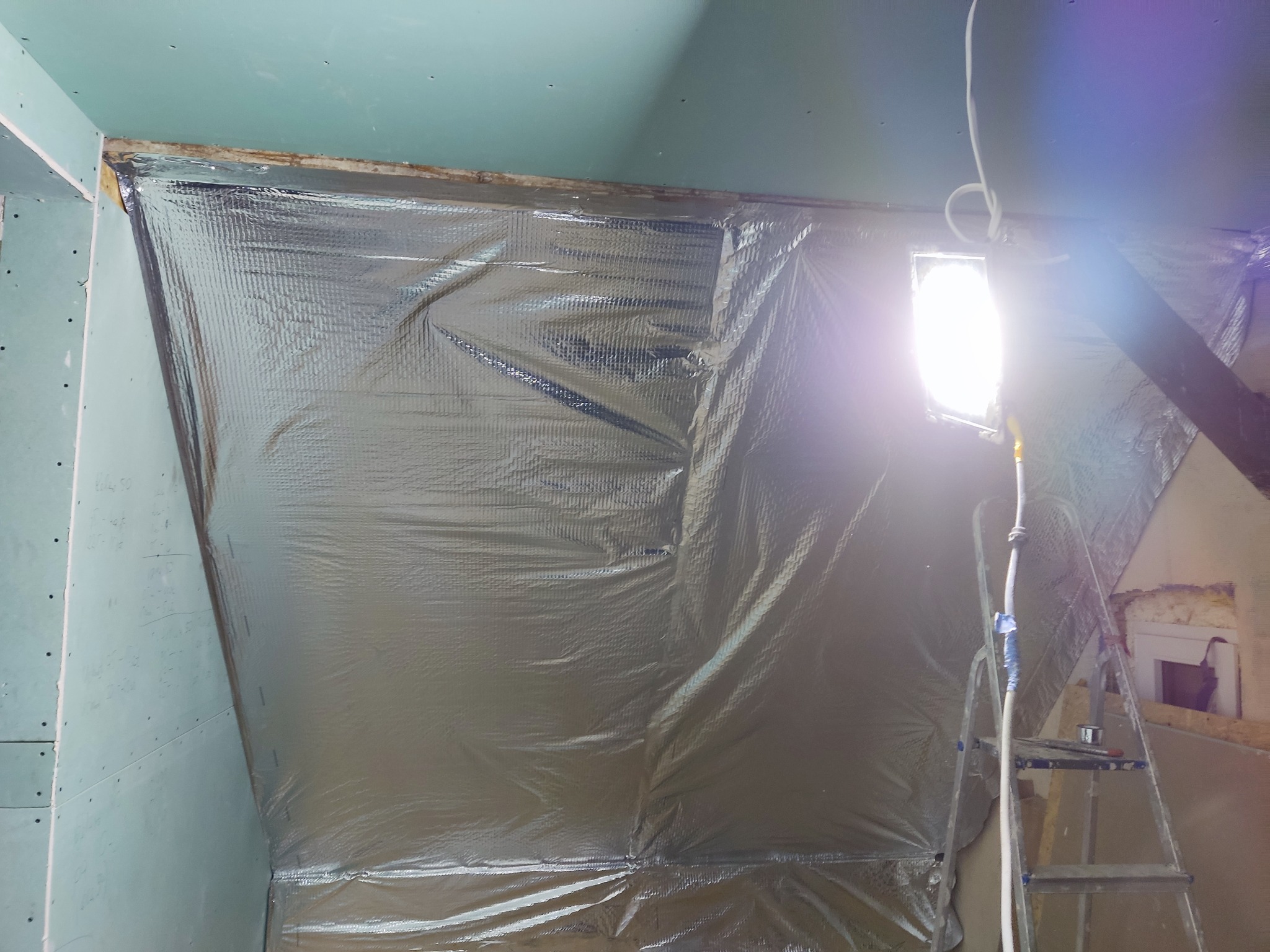 Montaż folii paroizolacyjnej na ścianie z płyt karton-gips podczas remontu poddasza, widoczna drabina i halogen oświetlający miejsce pracy.