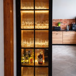 TENOMI Tomasz Wójcikiewicz - Czarna, przeszklona szafka z podświetleniem LED, wypełniona szkłem i butelkami alkoholu, w tle kuchnia z ceglaną ścianą i drewnianymi szafkami.