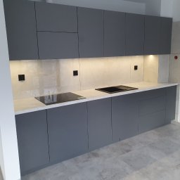 TENOMI Tomasz Wójcikiewicz - Nowoczesna kuchnia w odcieniach szarości z białym blatem i podświetleniem LED. Minimalistyczny design, płyta indukcyjna. Widok na fragment podłogi z szarych płytek.