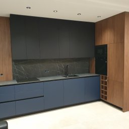 TENOMI Tomasz Wójcikiewicz - Nowoczesna kuchnia z granatowymi szafkami dolnymi, czarnymi górnymi i drewnianymi panelami. Uwagę przykuwa marmurowy backsplash i minimalistyczna armatura.