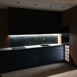 TENOMI Tomasz Wójcikiewicz - Nowoczesna kuchnia na wymiar z ciemnymi frontami i marmurowym blatem, oświetlona listwą LED, z drewnianymi elementami wykończenia.