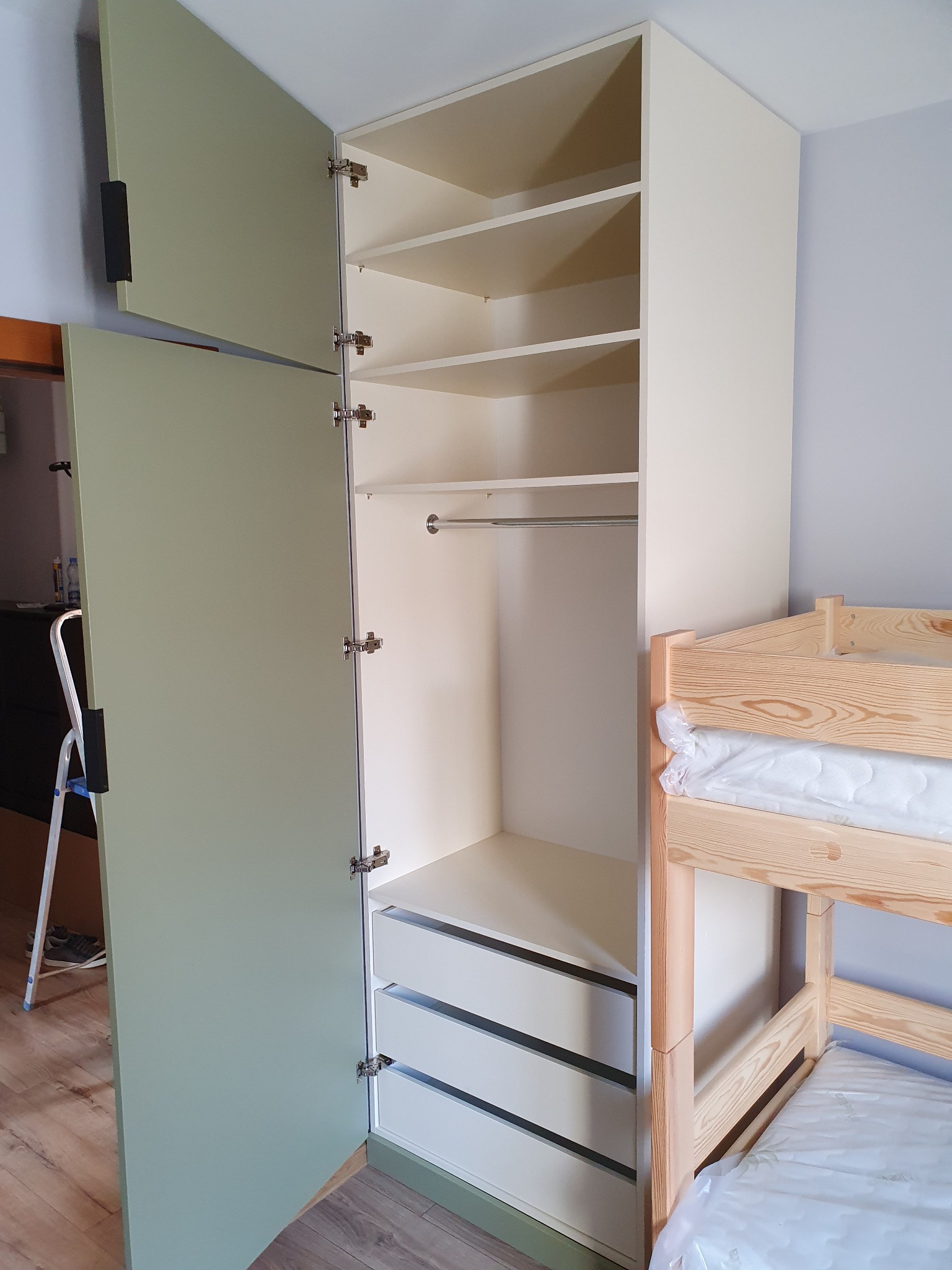 Szafa wnękowa z otwartymi drzwiami w kolorze pistacjowym, widoczne półki, szuflady i drążek na ubrania, obok łóżko piętrowe z surowego drewna.