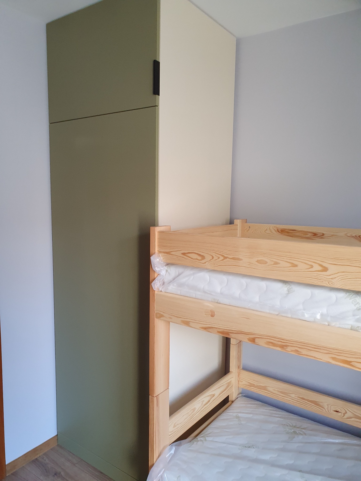 Wysoka, oliwkowa szafa wnękowa z czarnym uchwytem, przylegająca do drewnianego łóżka piętrowego z materacami w folii, na tle szarej ściany.