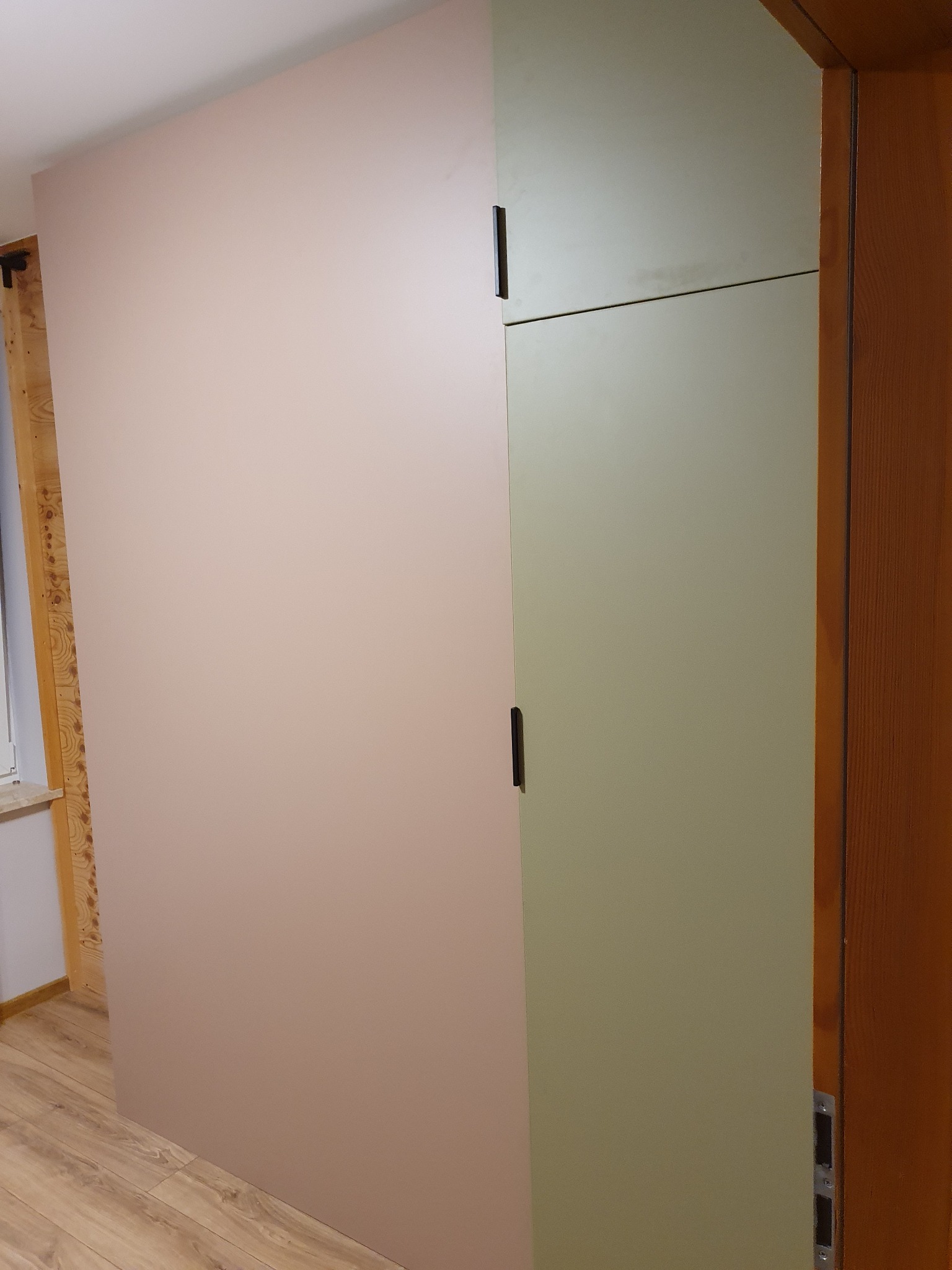 Wnętrze z dwukolorową szafą wnękową: pudrowy róż i oliwkowa zieleń, z minimalistycznymi, czarnymi uchwytami, widoczna drewniana rama okna i podłoga z paneli.