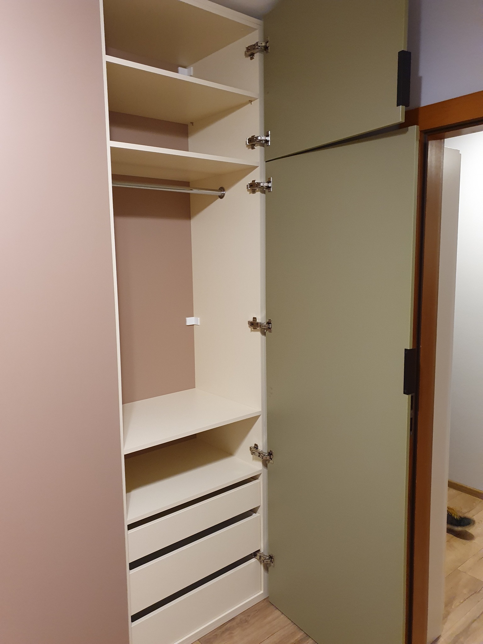 Otwarta szafa wnękowa z szufladami, półkami i drążkiem na ubrania, widok z boku na wnętrze z beżowymi ściankami i kremowymi frontami, brązowe wykończenie drzwi w tle.