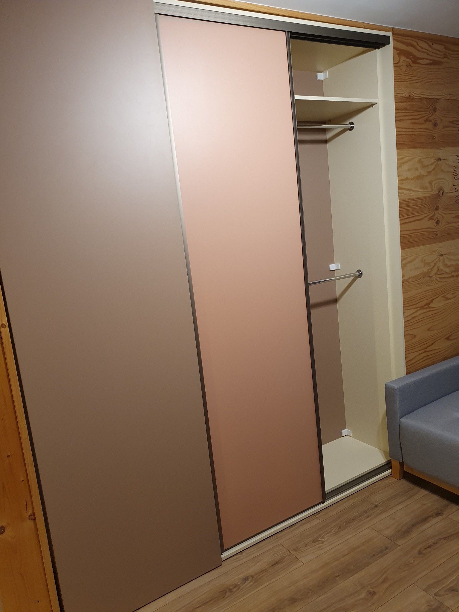 Wnętrze szafy wnękowej z przesuwnymi drzwiami w odcieniach brązu i różu, widoczny drążek na ubrania i półki, obok fragment sofy.