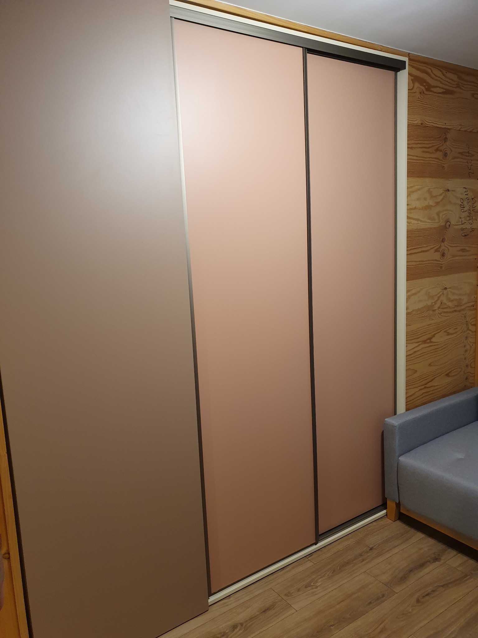 Trzydrzwiowa szafa wnękowa z przesuwnymi drzwiami w odcieniach beżu i różu, na tle drewnianej ściany i częściowo widocznej szarej sofy.