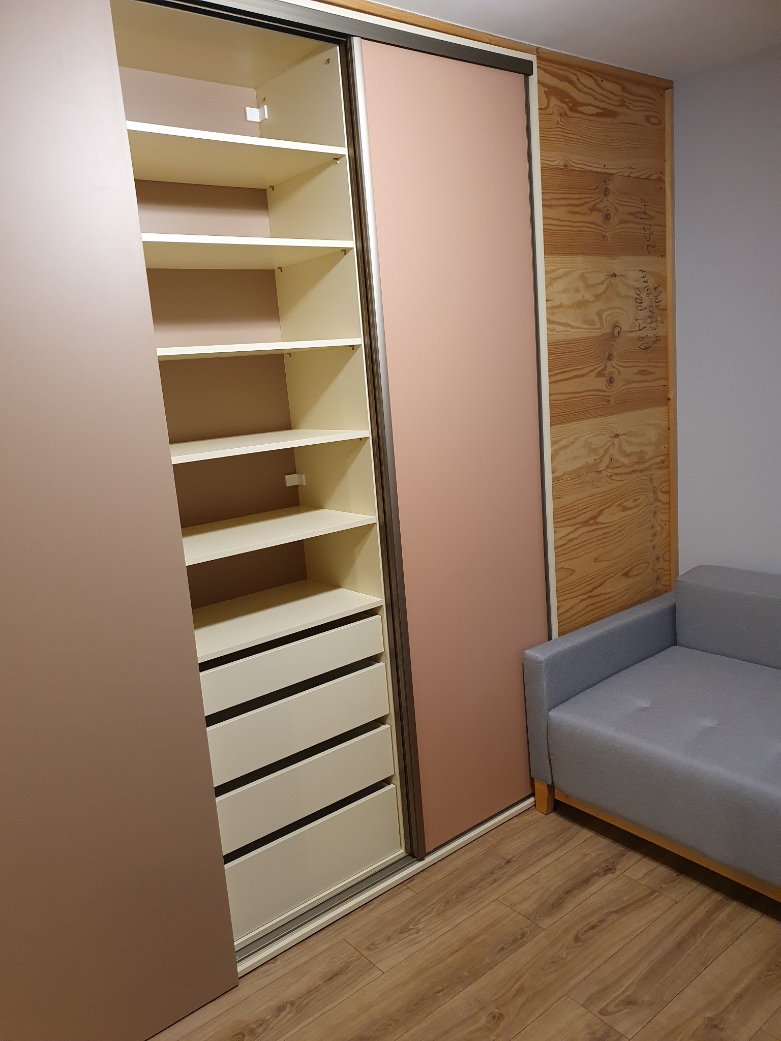 Wnętrze szafy wnękowej z przesuwnymi drzwiami w odcieniach beżu i różu, widoczne półki i szuflady, obok szara sofa, ściana z drewnianej boazerii z notatkami.