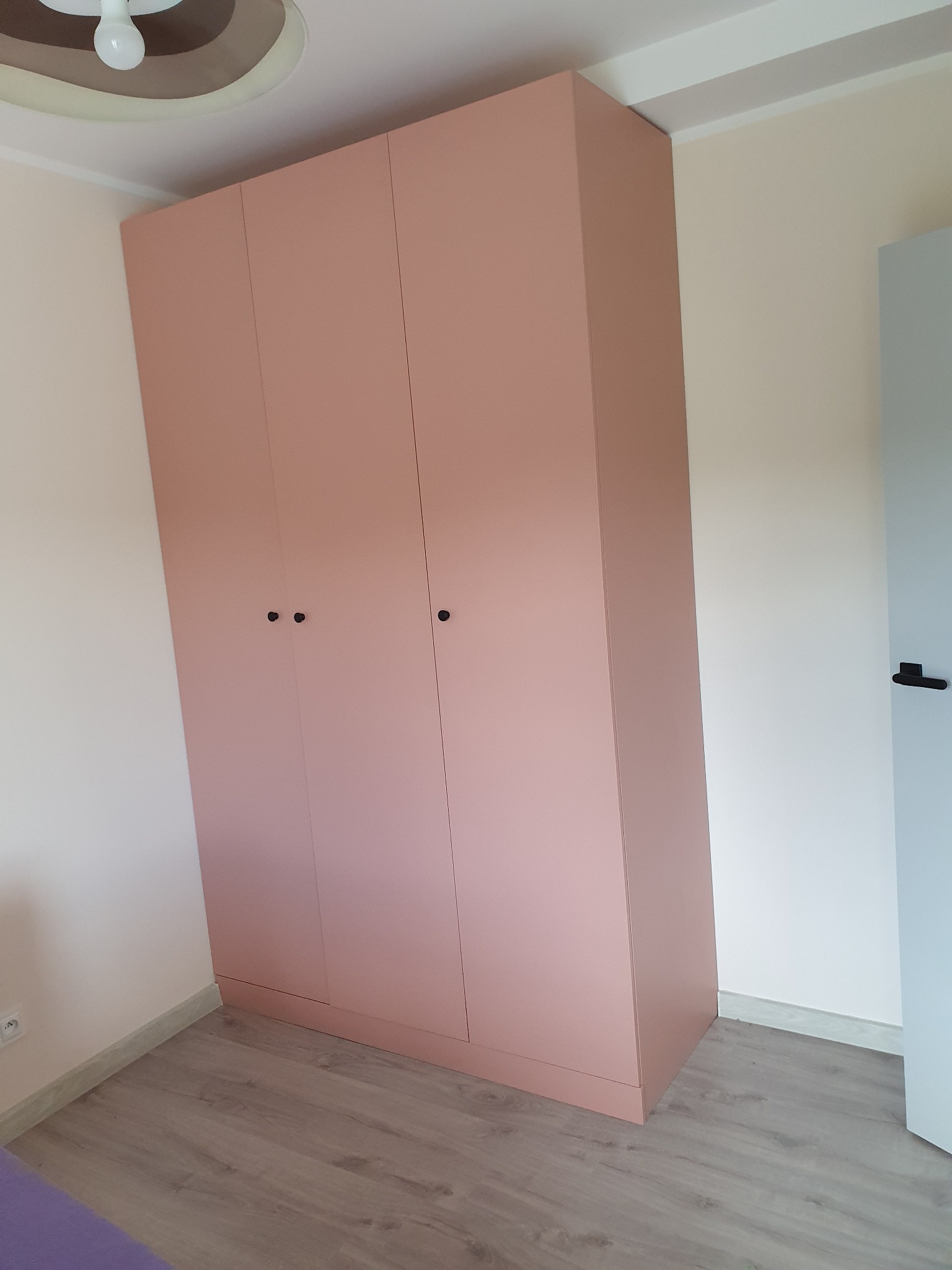 Różowa szafa wnękowa z trzema drzwiami na drewnopodobnej podłodze, widoczna lampa sufitowa i fragment ściany w odcieniach bieli.