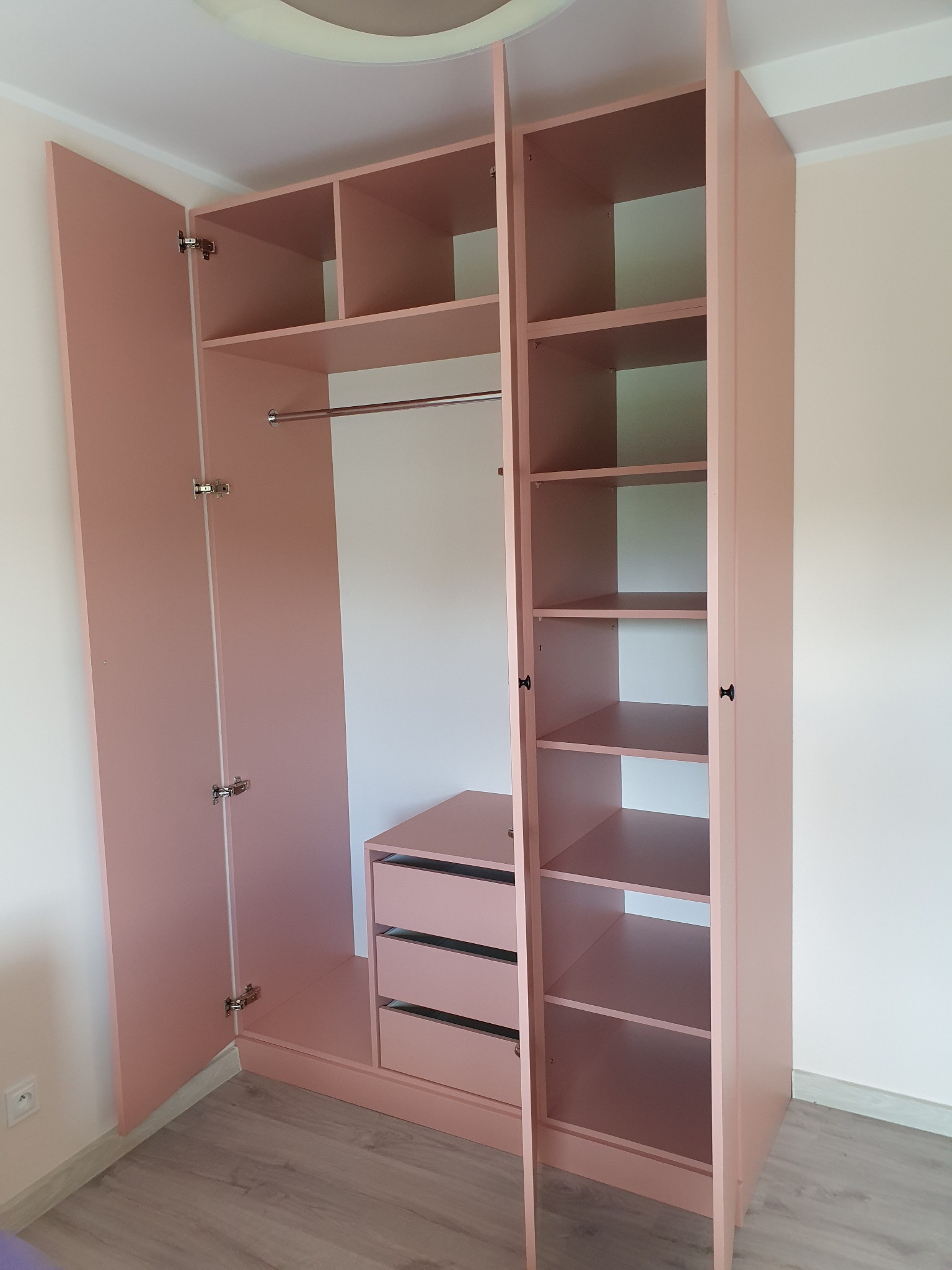 Różowa szafa wnękowa z otwartymi drzwiami, widoczne półki, szuflady i drążek na ubrania.