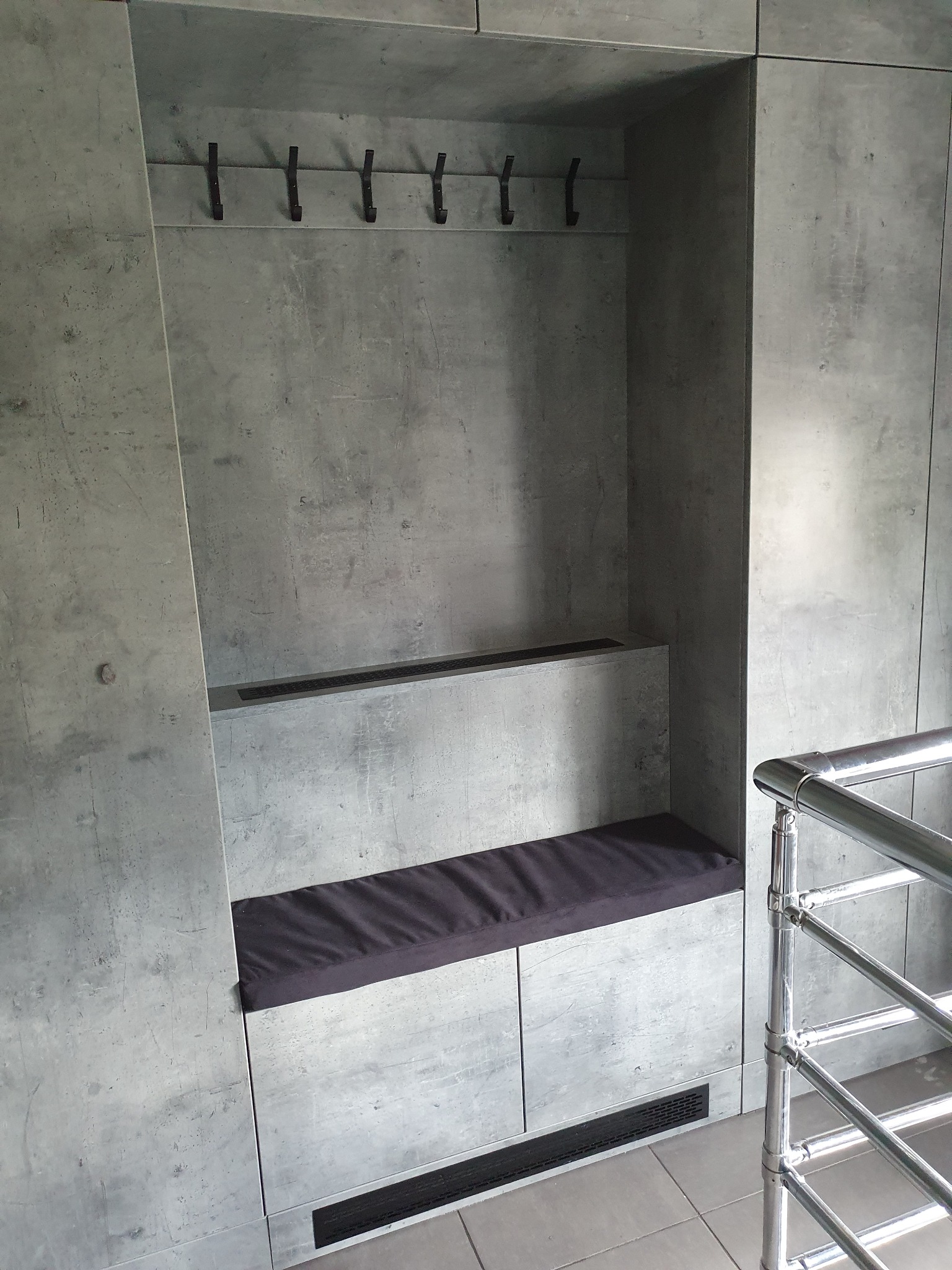 Szara zabudowa wnękowa z siedziskiem z ciemną poduszką i wieszakami na ubrania, imitacja betonu, nowoczesny design