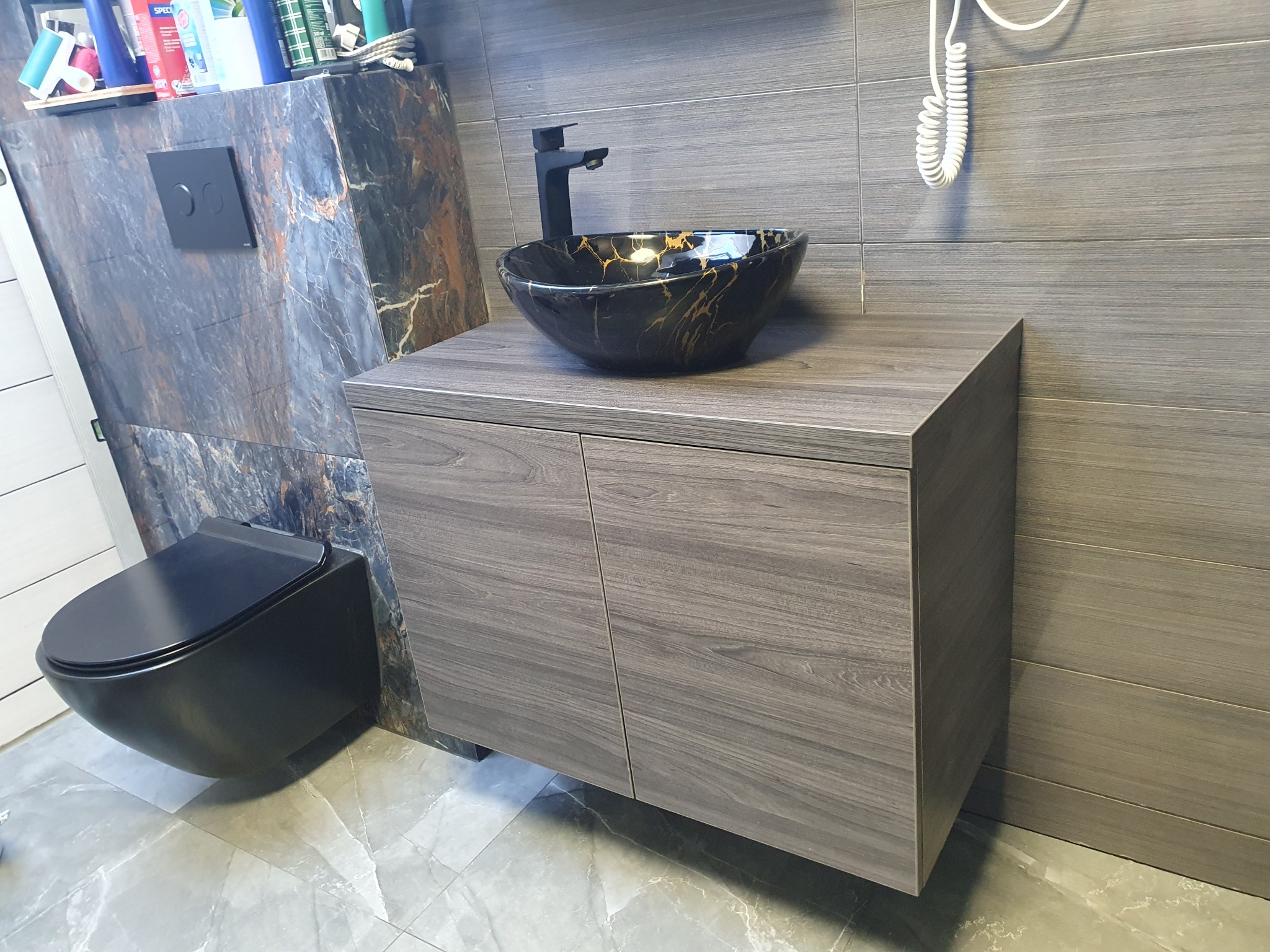 Nowoczesna łazienka z szarą szafką pod umywalkę, czarną umywalką z marmurkowym wzorem, wiszącą toaletą i szarymi płytkami imitującymi drewno na ścianie.