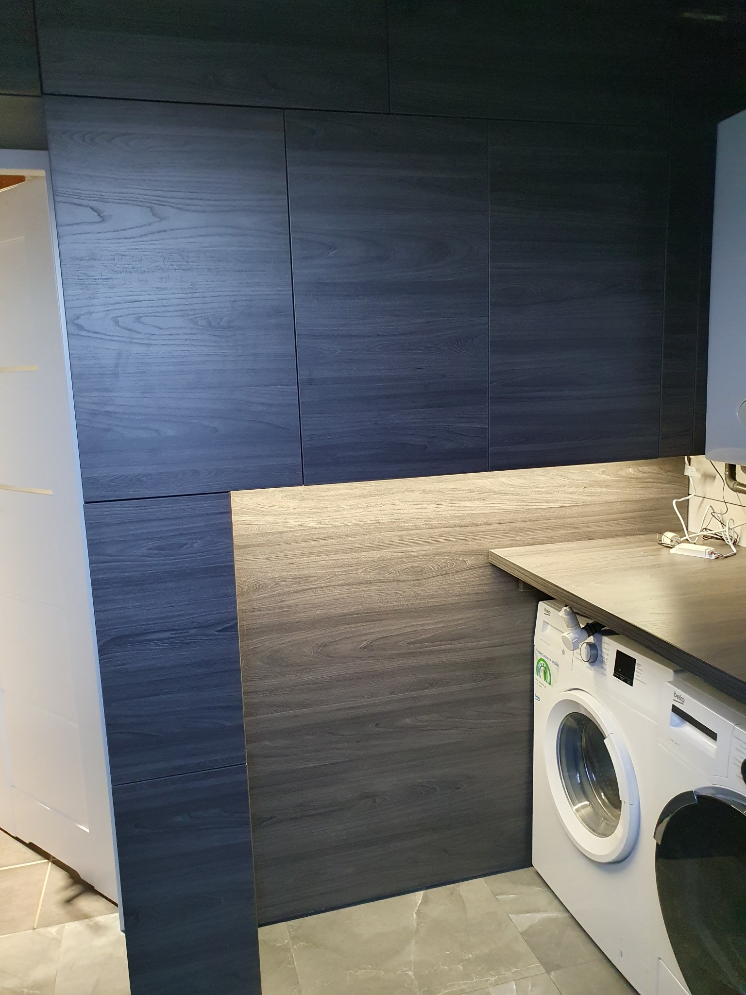 Ciemne, drewnopodobne szafki kuchenne sięgające sufitu, zintegrowane z blatem i pralką w nowoczesnej aranżacji wnętrza.