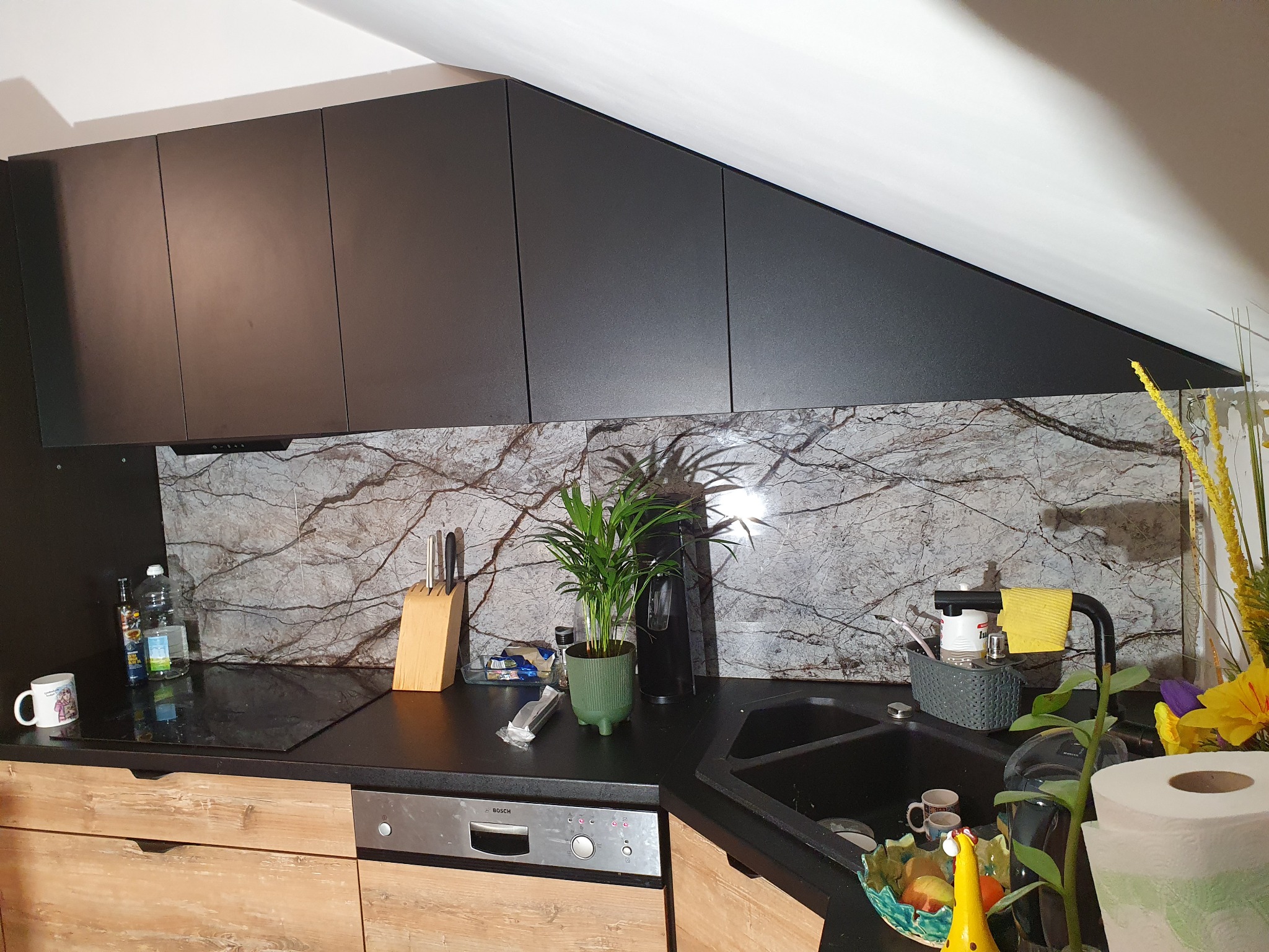Czarna zabudowa kuchenna pod skosem, drewnopodobne fronty dolnych szafek, marmurowy panel ścienny, zlewozmywak z drobnymi akcesoriami i kwiat doniczkowy.