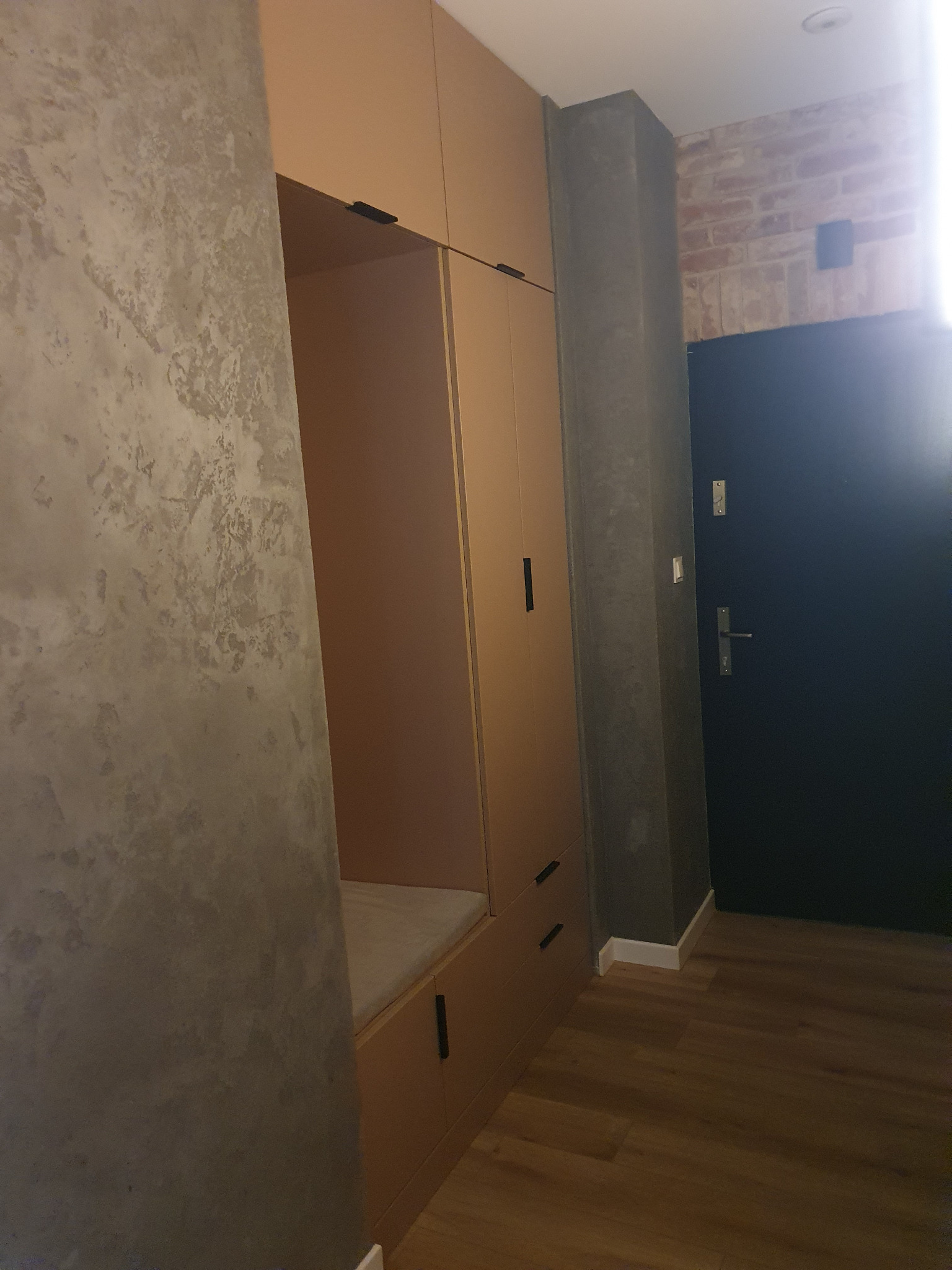 Beżowa szafa wnękowa z siedziskiem w przedpokoju, minimalistyczny design, czarne uchwyty, szara ściana z efektem betonu, drewniana podłoga, ciemne drzwi wejściowe.