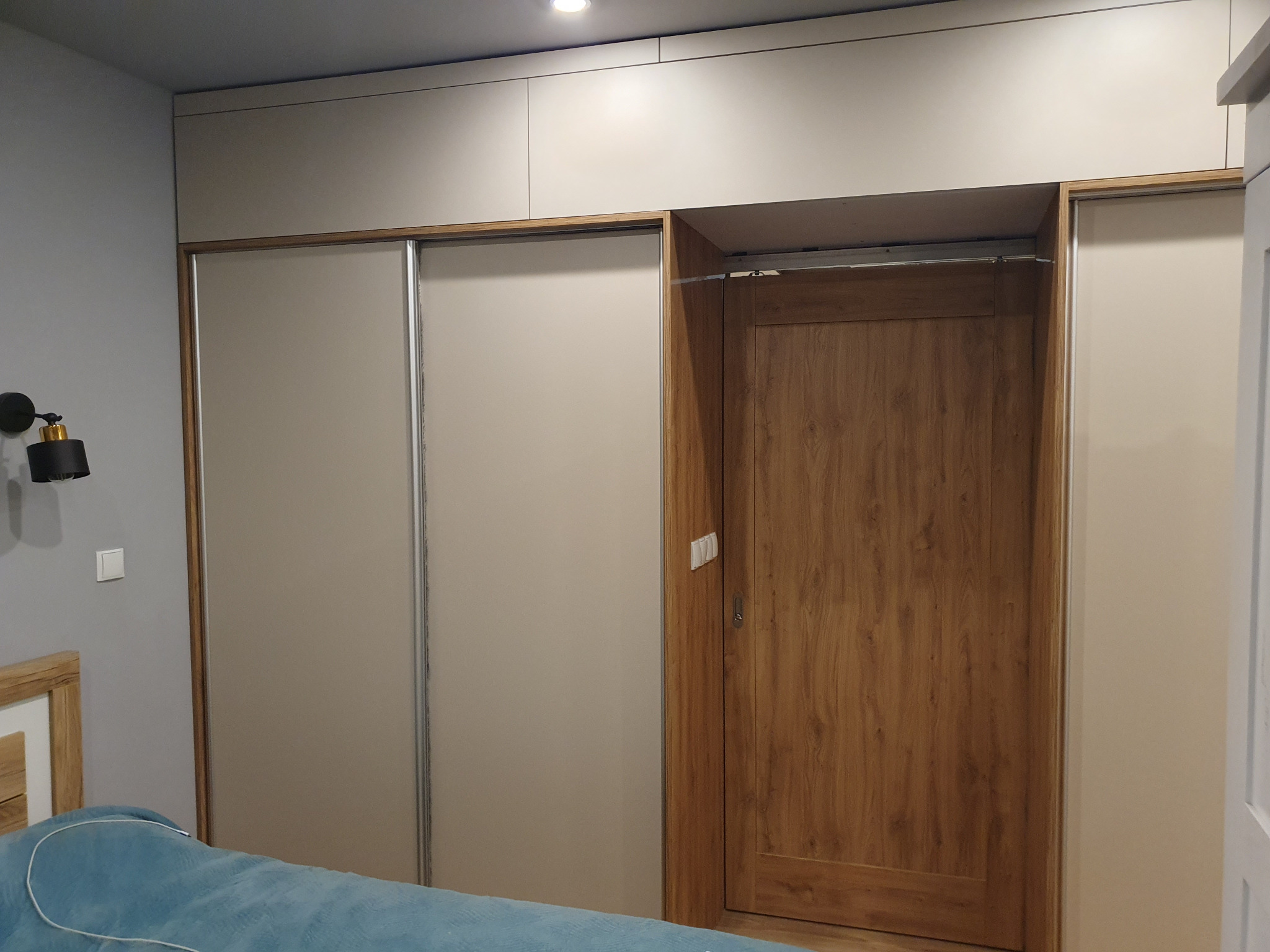 Szafa wnękowa z przesuwnymi drzwiami w odcieniach beżu i brązu, wkomponowana w ścianę sypialni, z widocznym fragmentem łóżka i lampki nocnej.