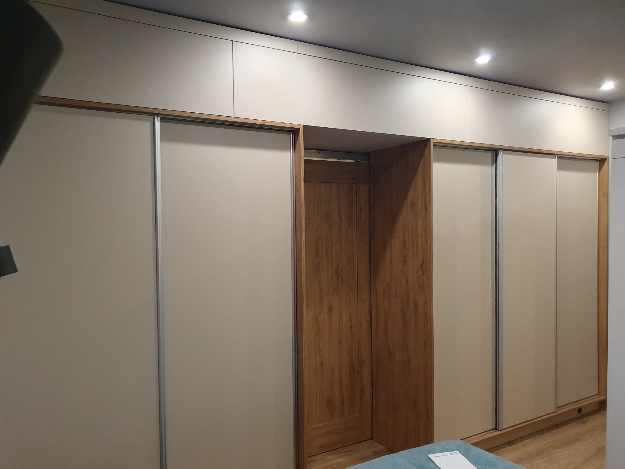 Beżowa szafa wnękowa z przesuwnymi drzwiami i drewnianym wykończeniem wnęki. Górne szafki do sufitu, oświetlenie punktowe. Minimalistyczny design.