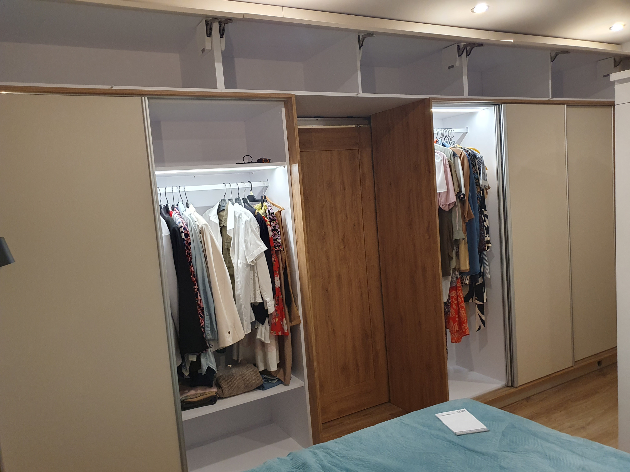 Beżowa szafa wnękowa z przesuwnymi drzwiami, oświetleniem LED wewnątrz i widoczną zawartością: ubrania na wieszakach. Częściowo otwarte drzwi z drewnianym wzorem.