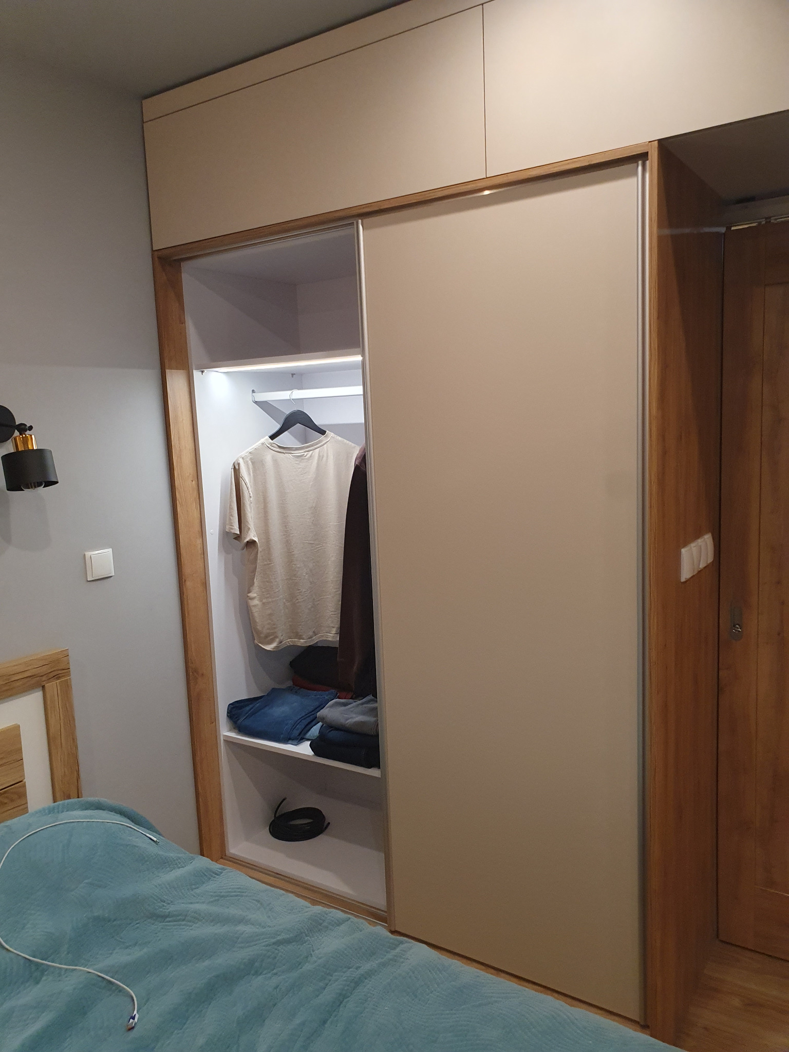 Szafa wnękowa z przesuwnymi drzwiami w sypialni, z widoczną zawartością półek i wieszaków. Jasne fronty, drewniane obramowanie, oświetlenie LED wewnątrz.