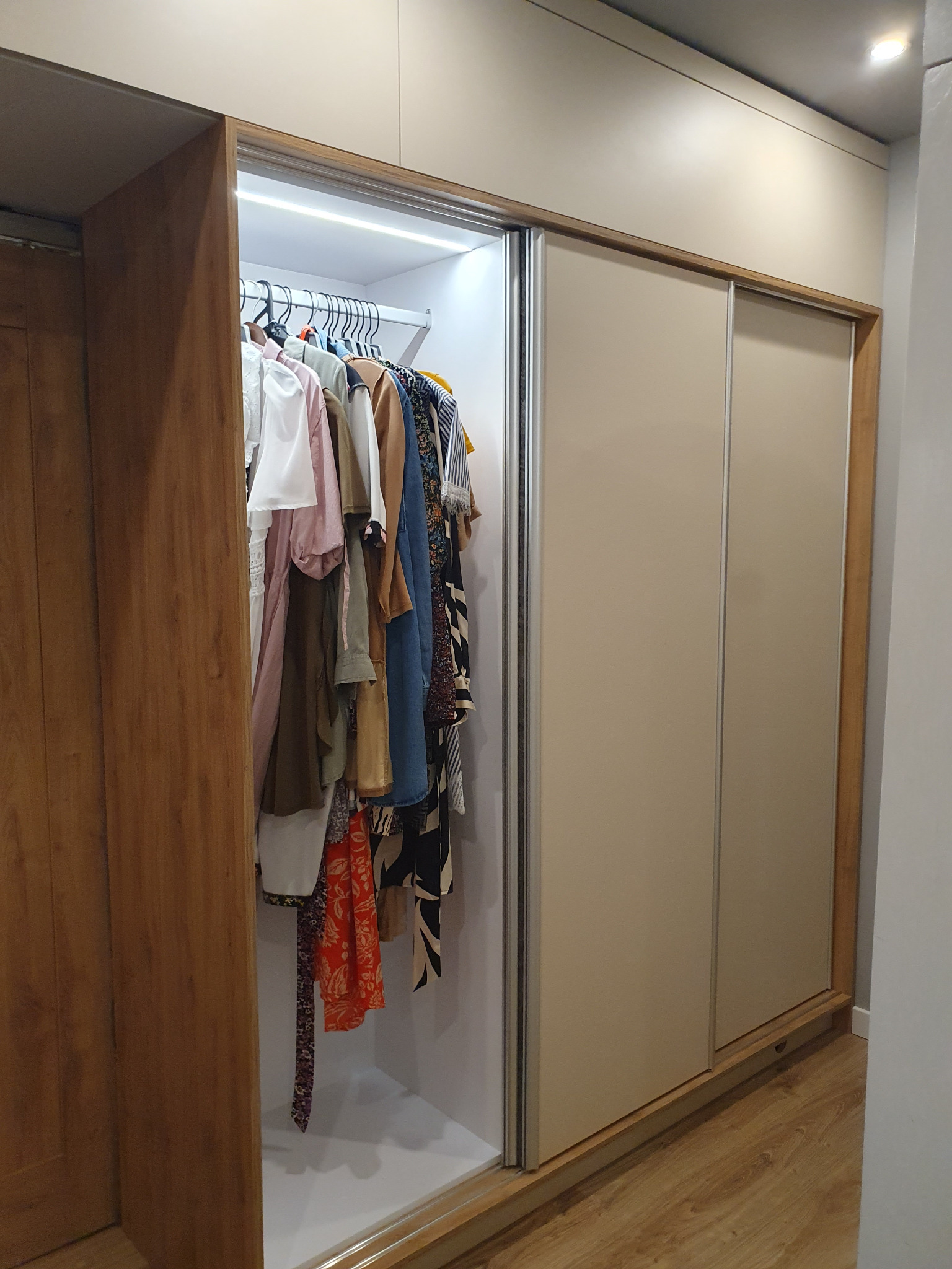 Beżowa szafa wnękowa z przesuwnymi drzwiami i drewnianym obramowaniem, oświetlona od wewnątrz, wypełniona ubraniami na wieszakach.