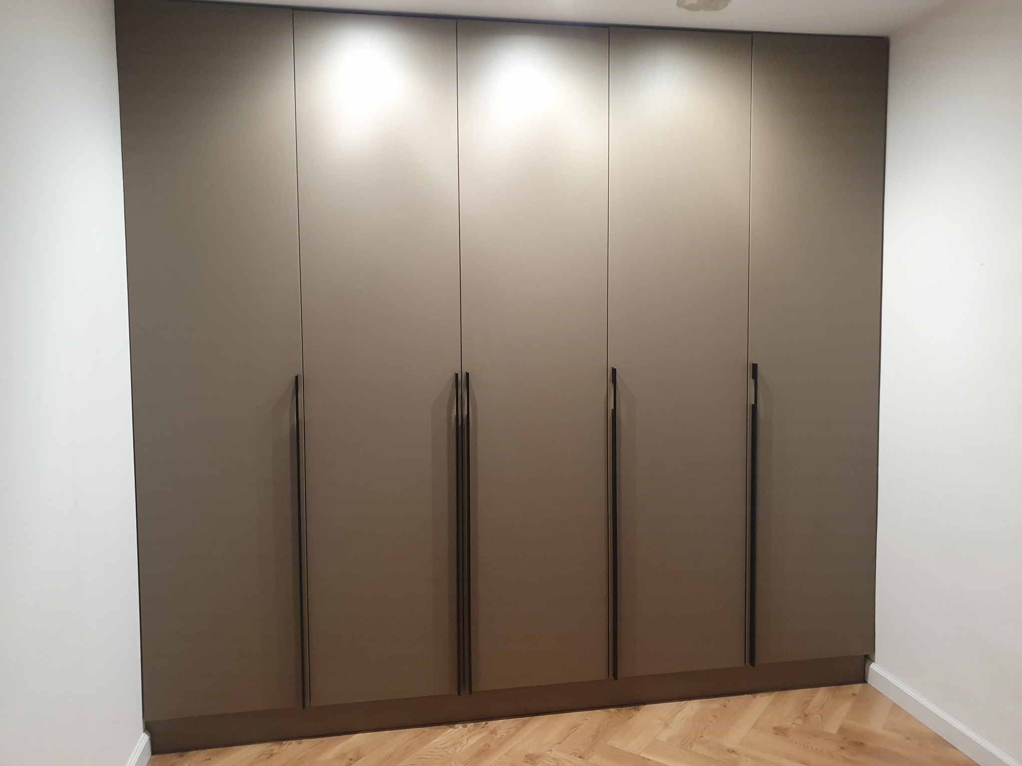 Beżowa szafa wnękowa z sześcioma drzwiami i długimi, pionowymi uchwytami, oświetlona od góry, na tle parkietu ułożonego w jodełkę.