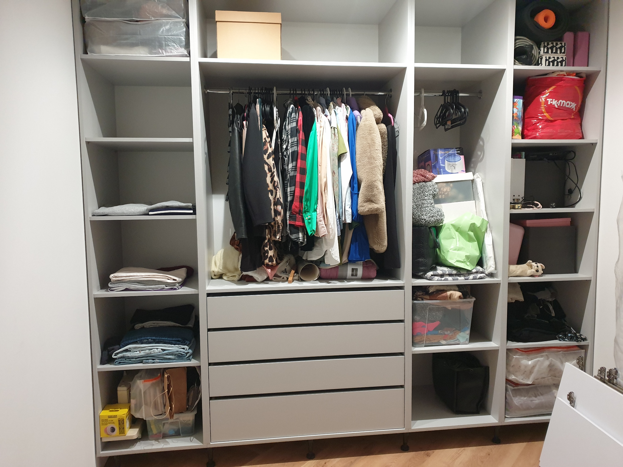 Jasnoszara szafa wnękowa z czterema szufladami, drążkiem na ubrania i półkami wypełnionymi ubraniami, pudełkami i akcesoriami. Widoczne detale wykończenia.