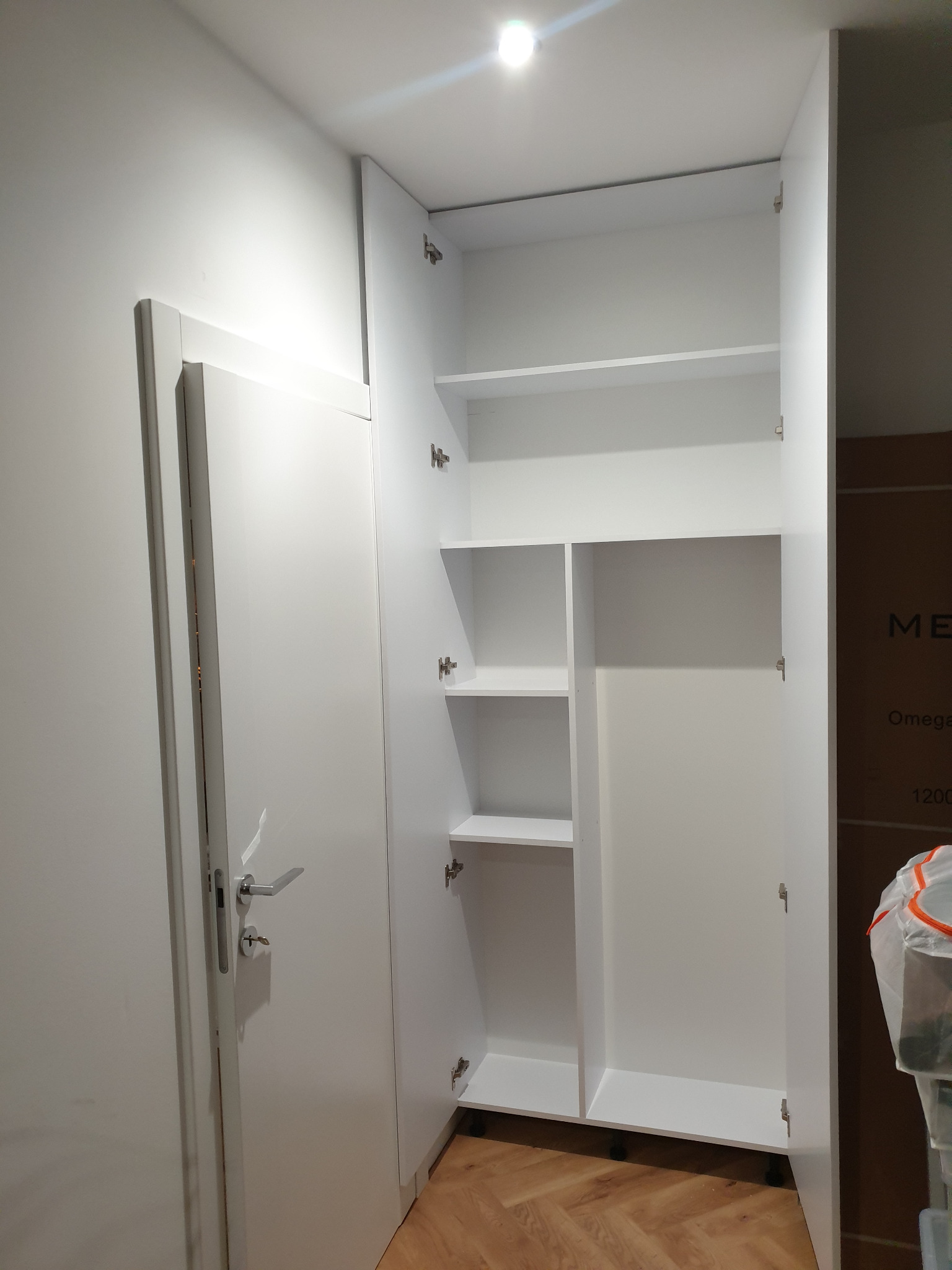 Biała szafa wnękowa z otwartymi drzwiami, widoczne półki i przestrzeń na wieszaki. Obok drzwi wejściowe, parkiet na podłodze. Minimalistyczny design.