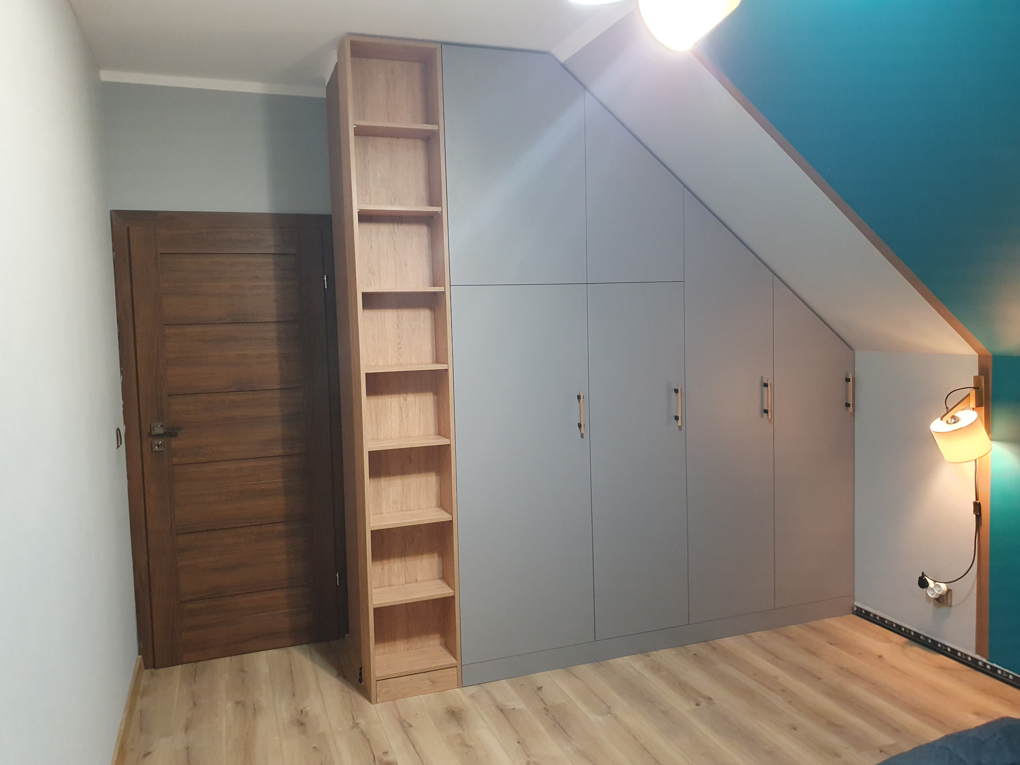 Szara szafa wnękowa z drewnianym regałem w pokoju na poddaszu, widok na drzwi i lampę ścienną. Minimalistyczny design, dopasowanie do skosu dachu.