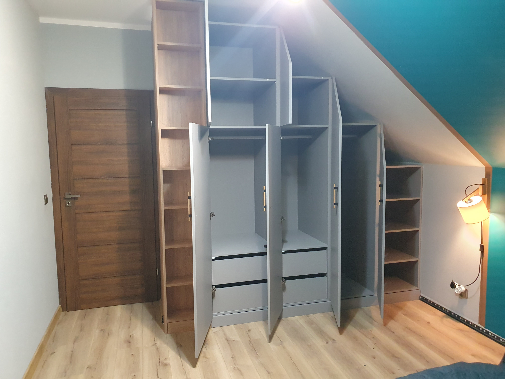 Szara szafa wnękowa z otwartymi drzwiami i szufladami, dopasowana do skosu dachu, z drewnianymi półkami po bokach i drewnianą podłogą.