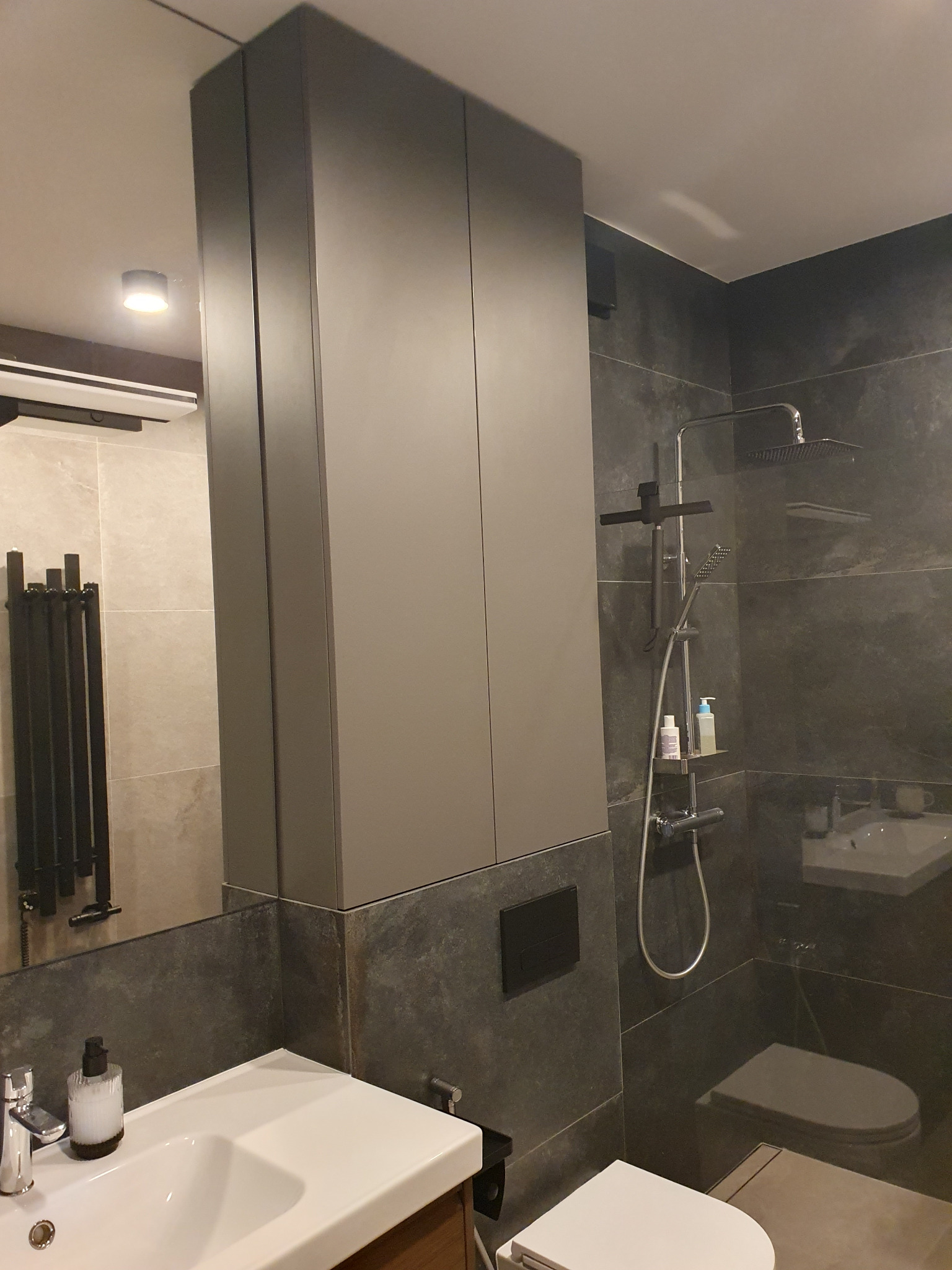 Nowoczesna łazienka z szarą szafką wnękową, umywalką i kabiną prysznicową z deszczownicą. Minimalistyczny design, ciemne płytki imitujące kamień.