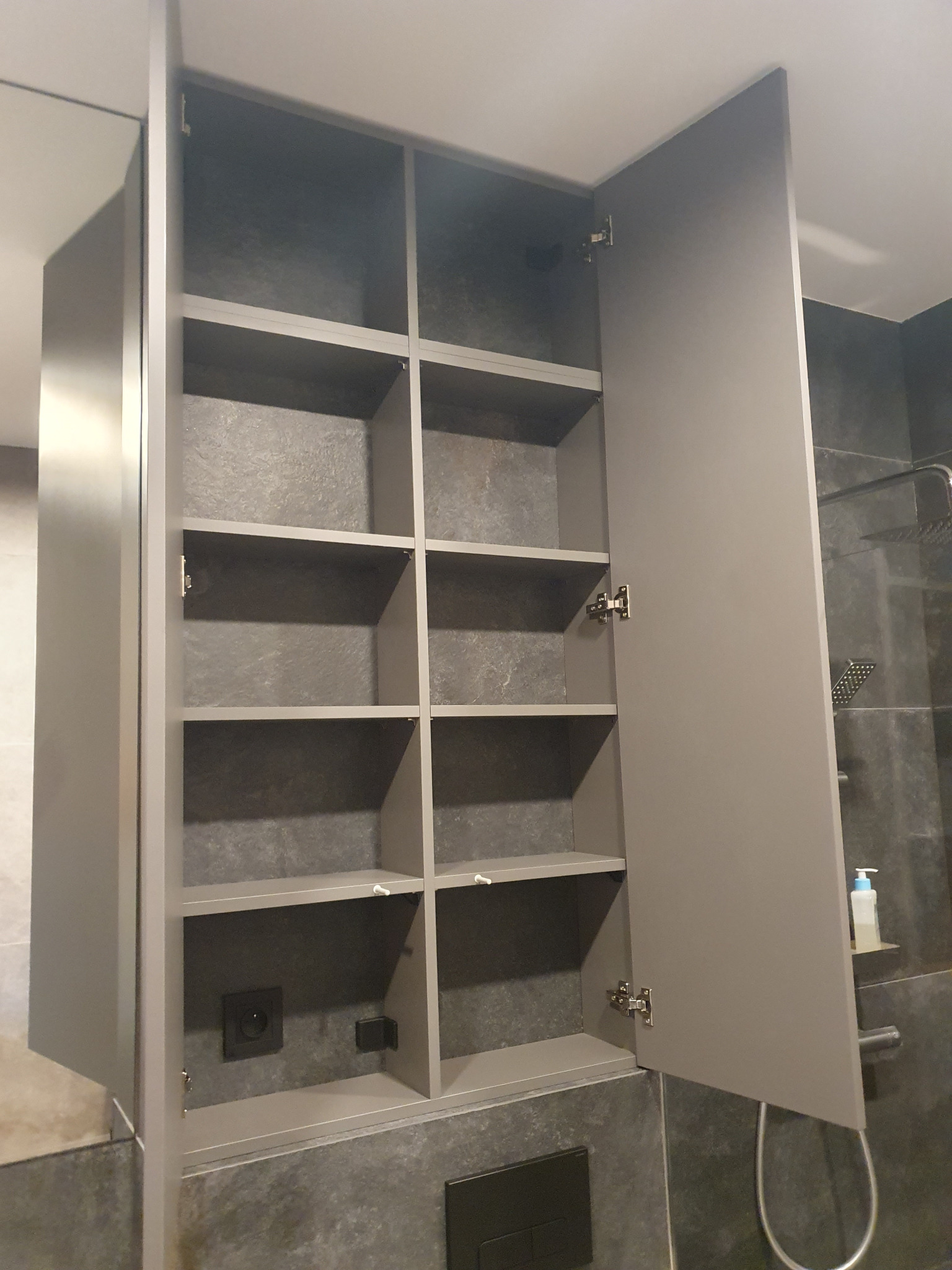 Szara szafka wnękowa z otwartymi drzwiami, widoczne półki i gniazdko elektryczne wewnątrz, na tle szarych płytek w łazience.