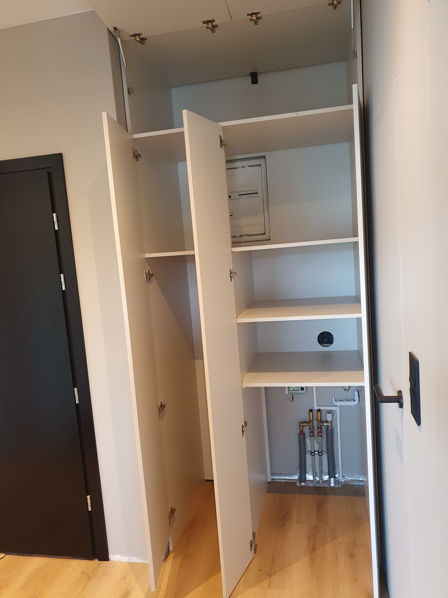 Biała szafa wnękowa z otwartymi drzwiami, ukazująca półki i instalacje. Widoczne zawiasy i mechanizmy otwierania. Minimalistyczny design, jasne wnętrze.