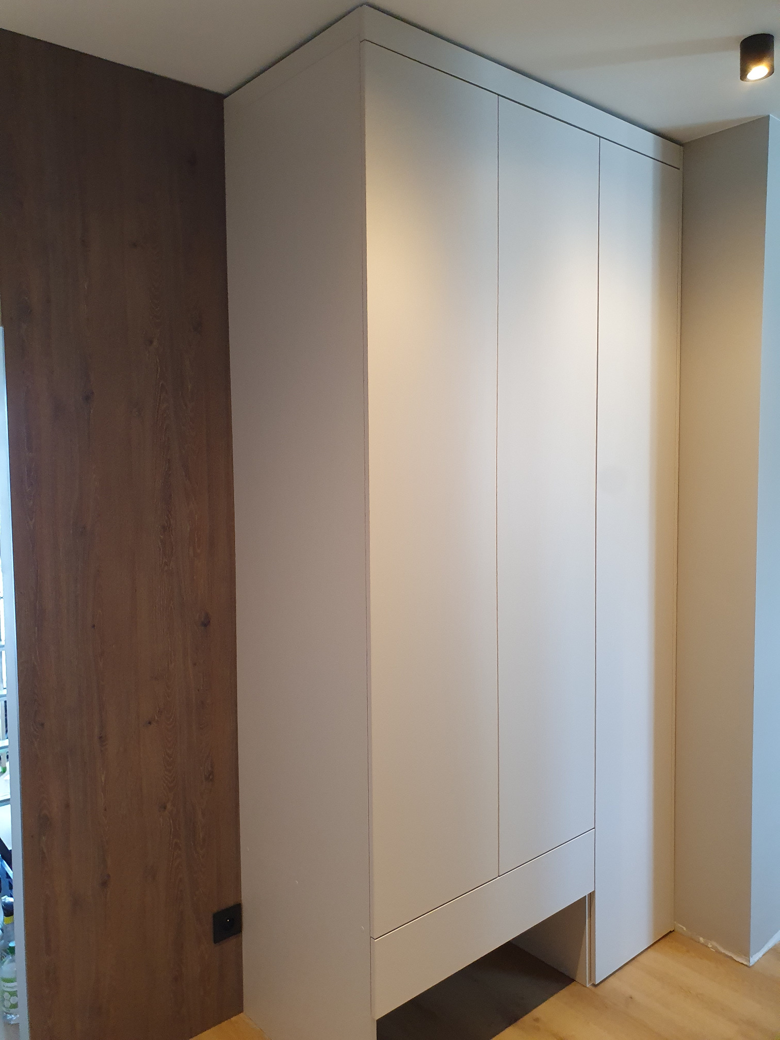 Jasnoszara, trzydrzwiowa szafa wnękowa z dolną przestrzenią, wkomponowana w narożnik pokoju z drewnianą ścianą i parkietem. Minimalistyczny design, oświetlenie sufitowe.