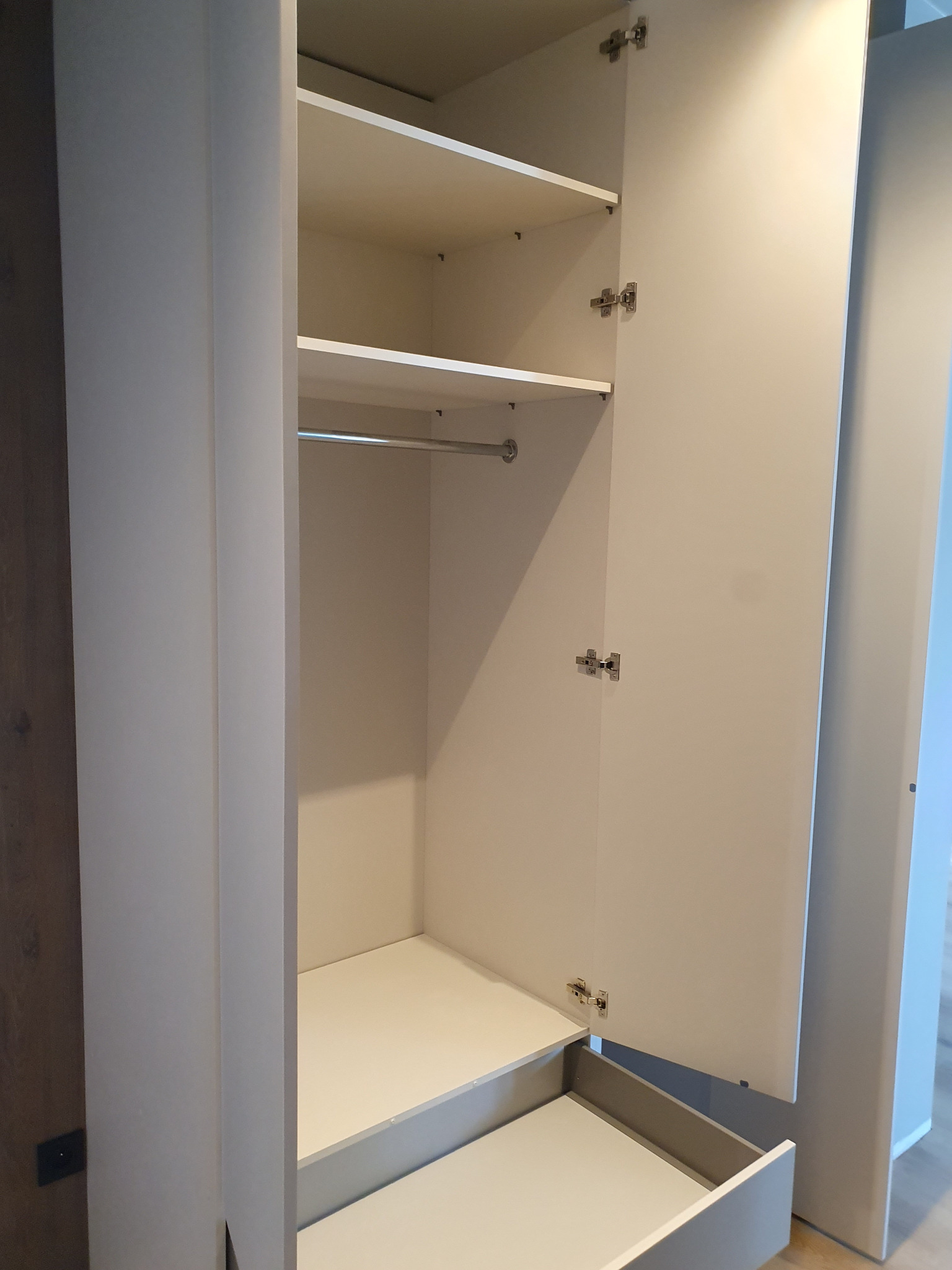 Otwarta, biała szafa wnękowa z wysuwaną szufladą i półkami, widoczne zawiasy, minimalistyczny design, oświetlenie dzienne.