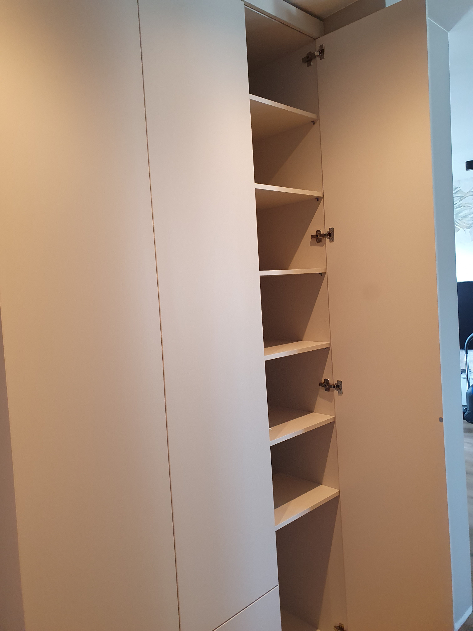 Otwarta, jasna szafa wnękowa z półkami, widoczne zawiasy. Minimalistyczny design, gładkie fronty. Ujęcie z dołu, podkreślające wysokość mebla.