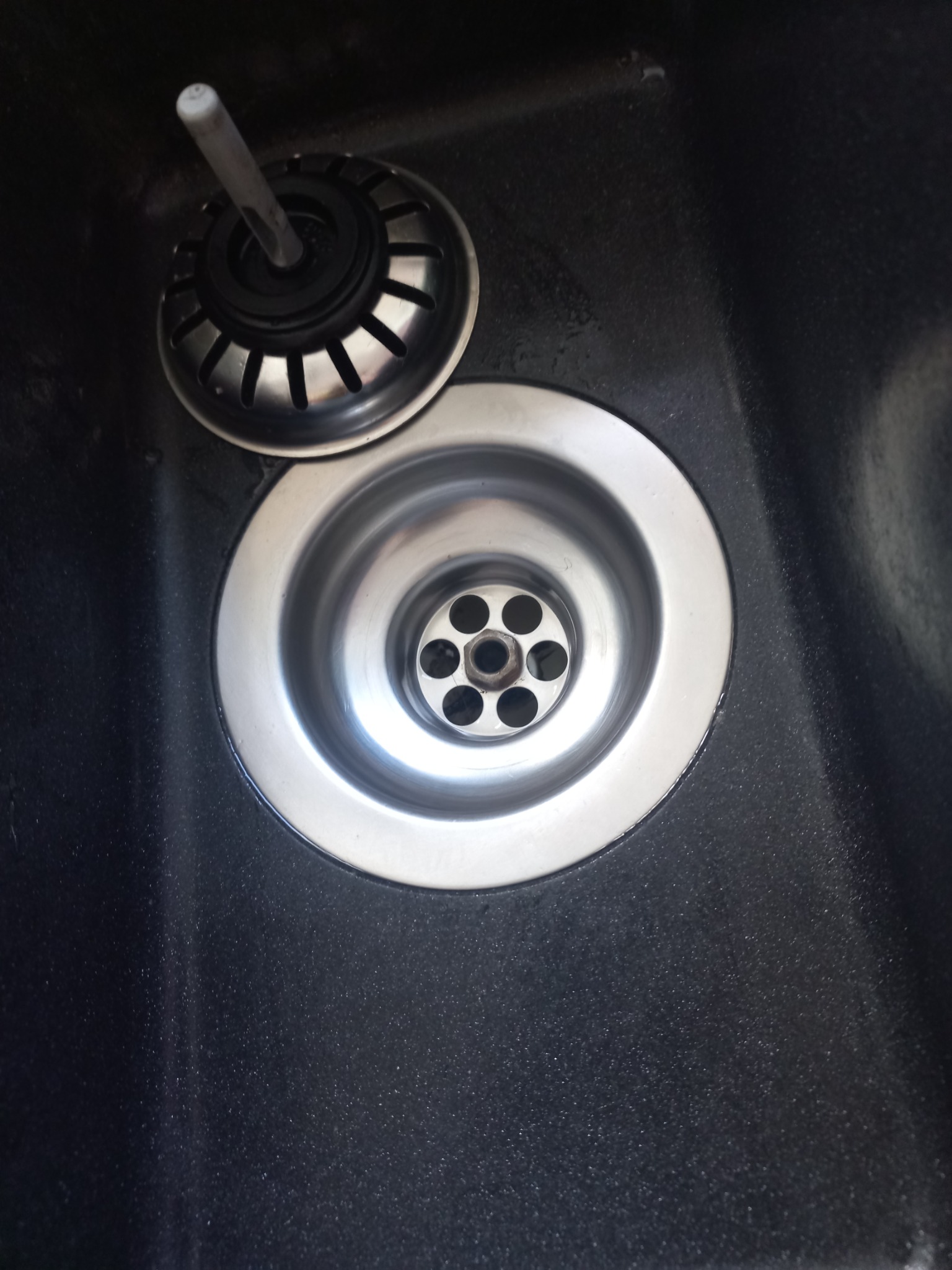 Zdemontowany odpływ zlewozmywaka z widocznymi elementami w ciemnym, ziarnistym zlewie.