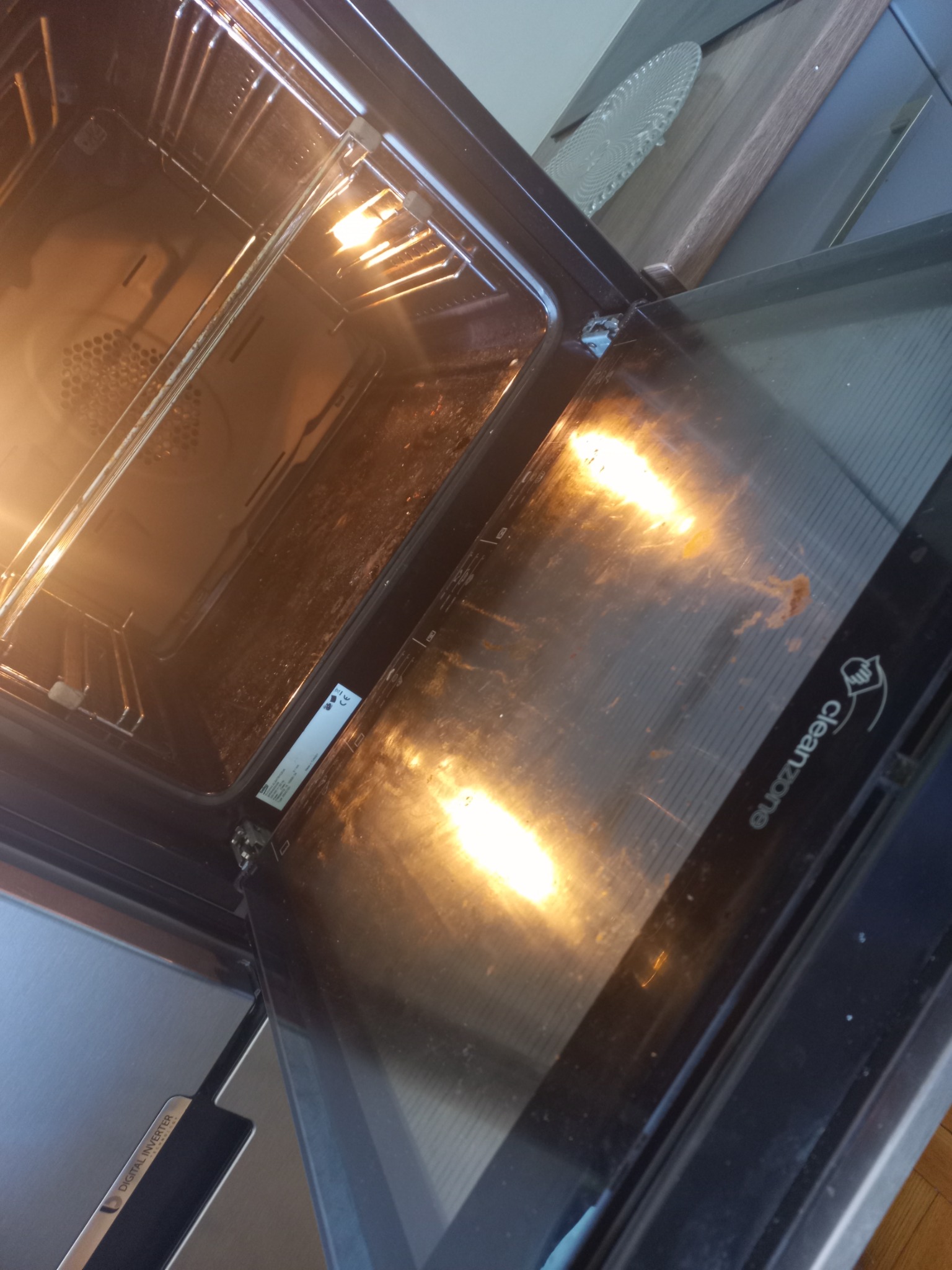 Wnętrze brudnego piekarnika z otwartymi drzwiami, widoczne przypalone resztki jedzenia na ściankach i szybie.